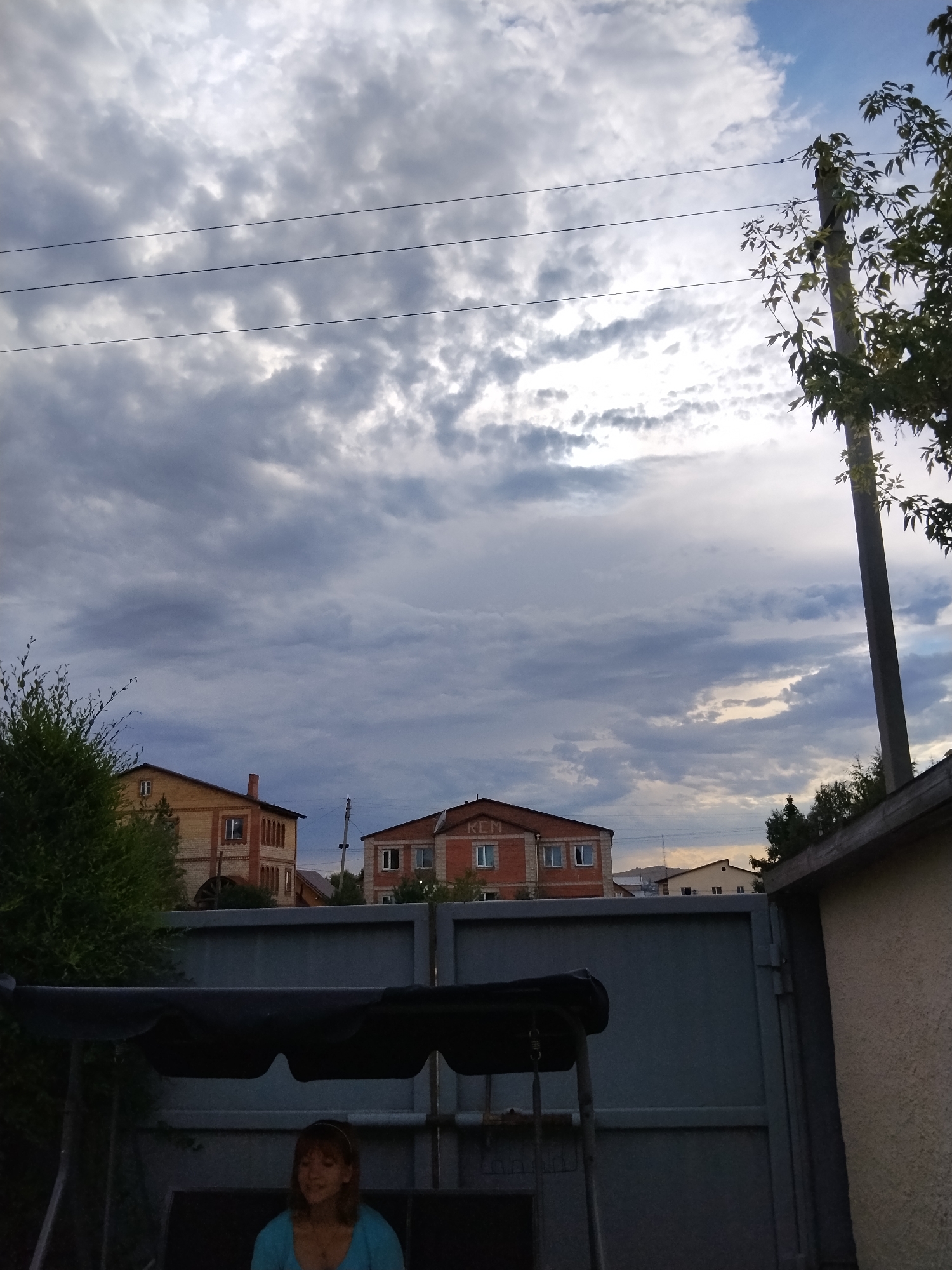 A medium shot captures a woman sitting on a swing set in a yard with a grey metal fence and a darker grey swing canopy. She has short, light brown hair and is wearing a light blue top and a silver necklace. The woman is looking down and to her left, her mouth slightly open. In the background, two brick buildings are visible under a cloudy sky. The building on the left is yellow and brick, and the building on the right is red brick with the letters "KCM" visible on its facade. A utility pole with power lines runs across the upper portion of the image. The scene appears to be in a residential area of Kokshetau, Kazakhstan.