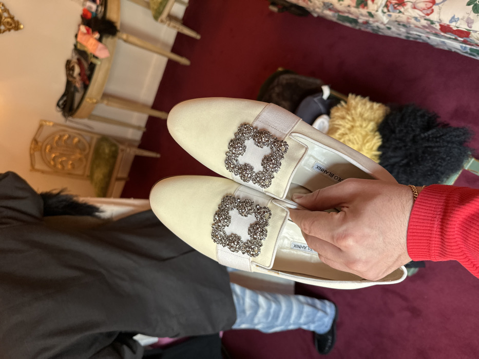 A pair of light cream Manolo Blahnik flat shoes, each featuring a square, crystal-studded buckle over a beige strap on the vamp, are held by a hand in the foreground. The hand wears a red long-sleeved top and a gold chain bracelet. The brand name "MANOLO BLAHNIK" is visible on the shoes' insoles. In the midground, a person is observed from behind, dressed in a dark jacket with visible fur trim, light blue patterned jeans, and black slip-on shoes. The scene takes place indoors on a deep red carpet. The background reveals a luxuriously appointed room, consistent with a high-end boutique or private dressing area in the city of London, United Kingdom. Visible objects include an ornate, gold-painted console table with miscellaneous items like socks scattered on its surface, and two matching gold-framed chairs with green upholstered seats.
