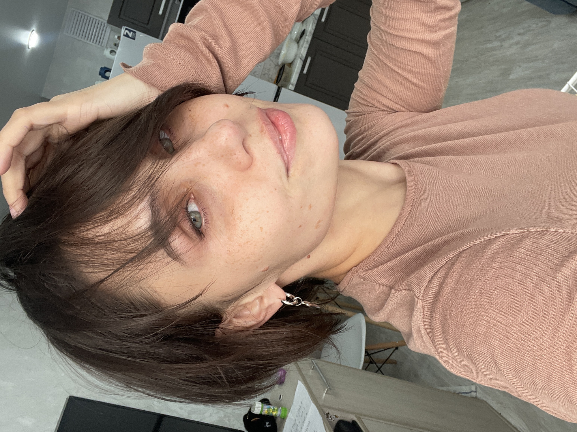 A close-up selfie depicts a young woman in an indoor setting in Izhevsk, Russia. She has fair skin with numerous freckles across her nose and cheeks, light-colored (likely grey or green) eyes, and full lips tinted with a natural or light pink shade. Her dark brown hair is styled casually, with strands falling across her forehead and face. She wears a long-sleeved, ribbed top in a blush pink or peachy hue, and a distinctive silver-toned earring, featuring interconnected hoops, is visible on her left ear. Her expression is calm and neutral as she looks directly towards the camera.
The background suggests a home environment, specifically a kitchen or living area. Visible elements include dark cabinetry, possibly a white countertop, a white appliance (likely a refrigerator or microwave) with a prominent black number "2" on its front, and a modern light fixture on the ceiling. Towards the bottom of the image, parts of a light-colored table or desk are visible, with papers (containing illegible handwritten text), a green-capped bottle, and a black object resting on it. A partial view of a white chair and a grey floor complete the domestic scene. The lighting indicates it is daytime.