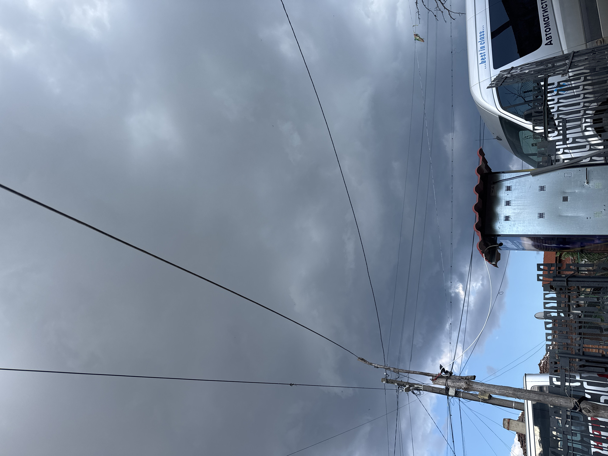 The image, taken in the city of Sredets, Bulgaria, depicts an overcast sky with heavy, dark clouds, suggesting impending rain. A power pole with numerous wires is prominently featured on the left side of the frame. In the foreground, a silver van with the text "...best in class..." and "Автомагист" is partially visible behind a black metal fence. To the left of the van, another vehicle and a corrugated metal structure with a red roof are present. The overall impression is of a somewhat utilitarian and possibly working-class urban environment under a dramatic sky.