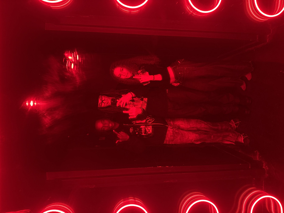 The image displays three individuals positioned centrally within a narrow, dark passage intensely illuminated by red light, located in the city of Paris, France. On the left, a male with dark skin and dark hair, wearing a dark zip-up hoodie over a graphic t-shirt and light-colored baggy trousers, raises his left hand with the middle finger extended towards the camera. In the center, a male wearing glasses, a dark beanie, and a dark graphic long-sleeved shirt with long dark hair, holds a smartphone in his right hand as if taking a photo, while also extending his left middle finger towards the camera. On the right, a female with long dark wavy hair, wearing a dark cropped long-sleeved top exposing her midriff and dark jeans, raises her left hand with the middle finger extended. The environment features dark walls and ceiling, with vertical arrays of circular red neon lights lining the left and right sides of the passage, and additional red light sources visible on the ceiling, creating a vibrant and confined atmosphere. All three subjects directly face the camera, collectively displaying a defiant gesture.