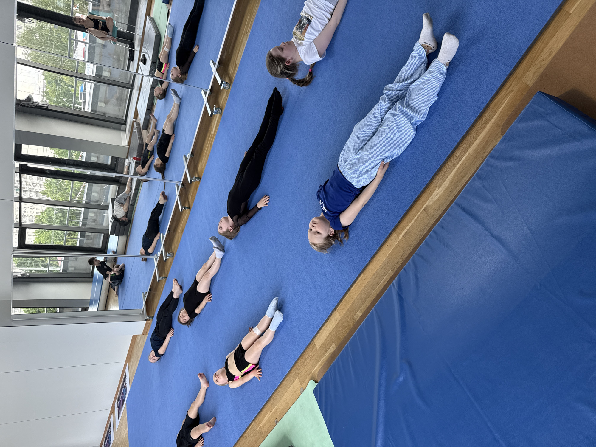 This image captures a group of young girls engaged in a training session, likely gymnastics or ballet, within an indoor studio setting in Khimki, Russia. The scene appears to be a moment of rest or stretching during their practice.
The main subjects are several girls, ranging in age from young children to pre-teens, most of whom are lying on their backs on a large, bright blue mat that covers a significant portion of the floor. Their positions suggest a relaxed or a specific stretching exercise. Their attire varies, including black athletic wear or leotards, some with colorful accents, and one girl prominently featured in light blue striped pants and a dark blue t-shirt. Another girl wears a white graphic t-shirt. Many have socks on, while others are barefoot. Their expressions are calm and focused.
The studio environment is characterized by a strip of light wooden flooring that runs alongside the blue mat. Beyond this, a tall mirrored wall extends across the left side of the frame, reflecting additional girls in various poses, some near horizontal bars (ballet barres). The reflections in the mirror also reveal large windows that look out onto an urban landscape, showing buildings, trees, and bright daylight, indicating it is daytime. A second, darker blue mat, possibly a thicker crash mat, is partially visible on the right side of the image, contrasting with the wooden floor. The overall atmosphere is one of a structured yet serene training environment. No legible text is visible in the image.