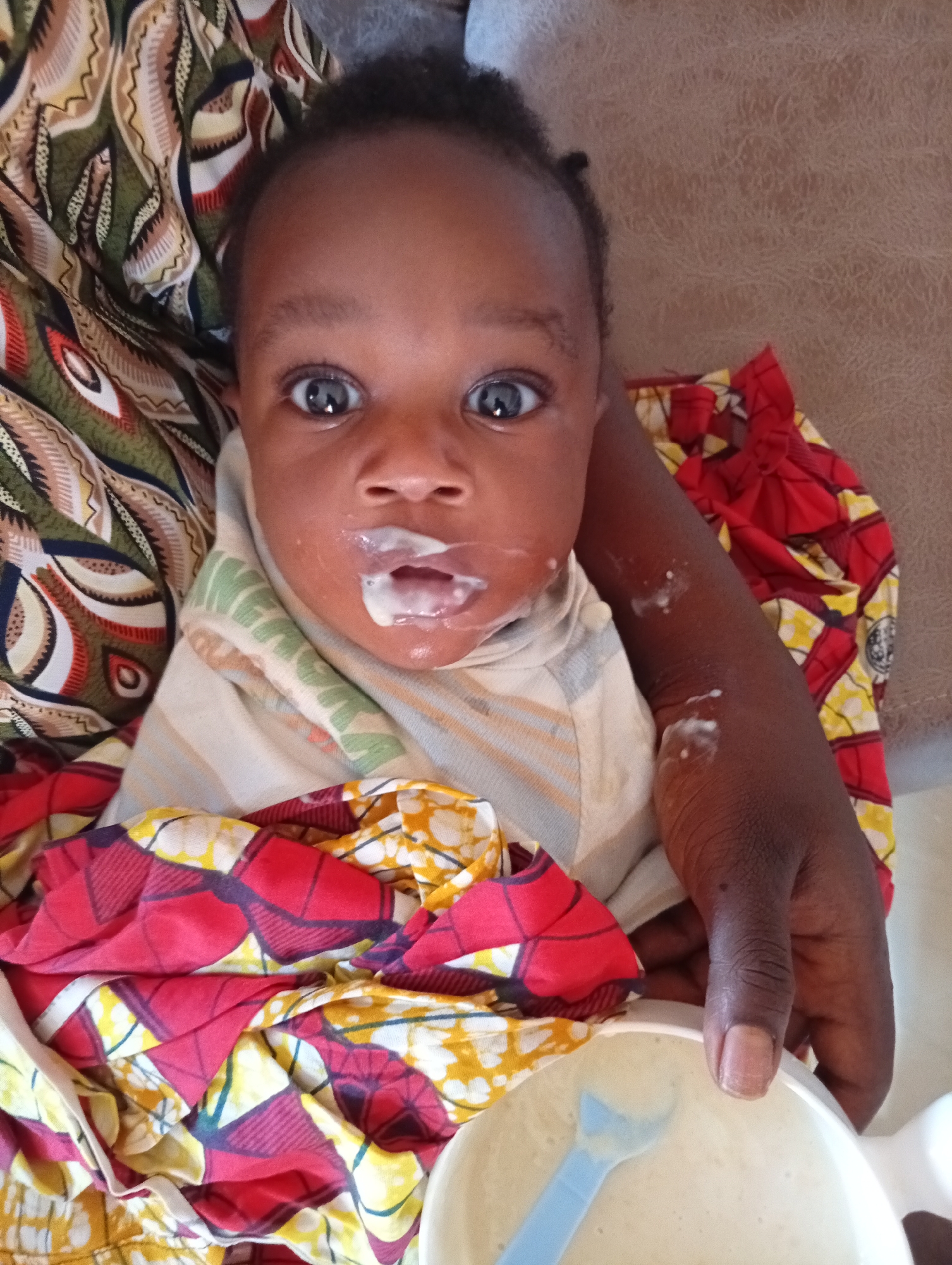 In Gura Top, Nigeria, a close-up, overhead shot shows a dark-skinned baby with wide, curious eyes. The baby's face and mouth are smeared with a white substance, likely food, and a small amount of the same substance is visible on its chin and arm. The baby is dressed in a light-colored garment with horizontal stripes and is swaddled in colorful, patterned fabrics featuring red, yellow, and green designs. A white bowl with a blue spoon rests in the foreground, partially filled with a pale, creamy substance. The background consists of a textured, light brown surface, possibly a sofa or bed. The lighting is natural, suggesting an indoor setting.