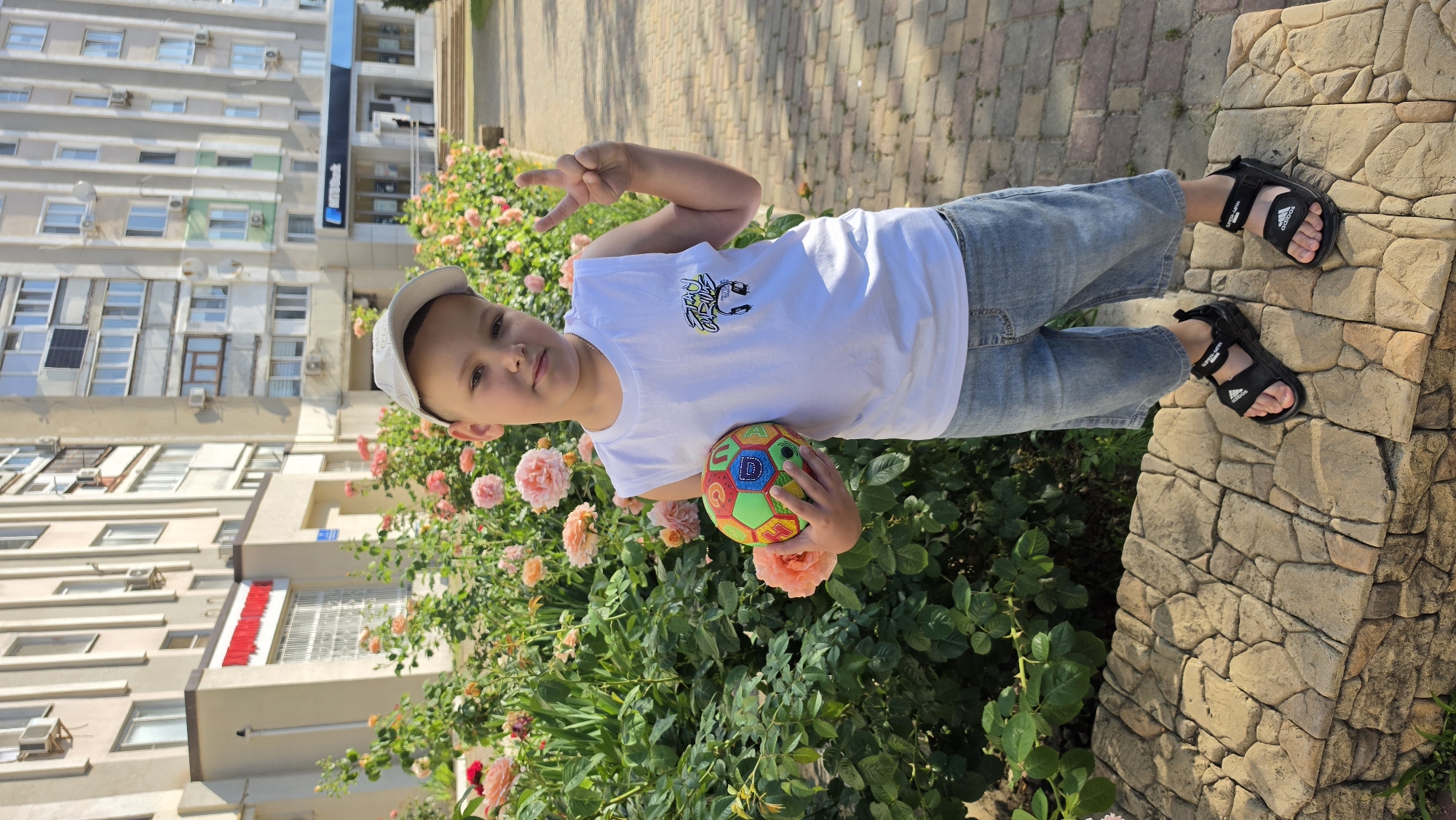 A young boy stands in front of a rose bush, holding a colorful ball and making a peace sign with his hand. He is wearing a white sleeveless t-shirt, denim shorts, and black sandals. In the background, a multi-story building with windows and air conditioning units is visible, along with a paved walkway and a stone bench. The scene appears to be outdoors in a city setting.