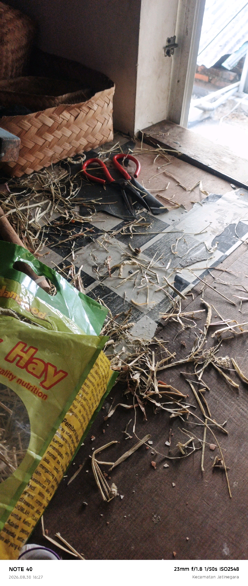The image is an overhead shot of a wooden surface covered with scattered dry grass and straw. A woven basket, a pair of red-handled scissors, and a green plastic bag with text in yellow and white are visible. The plastic bag appears to be for pet food or bedding, with the word "Hay" prominently displayed in yellow lettering. The scene suggests an indoor environment near a window, with some natural light entering. The bottom of the image contains a timestamp and location information. Specifically, the text at the bottom reads "NOTE 40 2026.08.30 16:27 23mm f/1.8 1/50s ISO2548 Kecamatan Jatinegara." This indicates the photograph was taken on August 30, 2026, at 16:27, with camera settings of 23mm focal length, 1/50s shutter speed, and ISO 2548. The location is identified as Kecamatan Jatinegara, which is a subdistrict within East Jakarta, Indonesia.