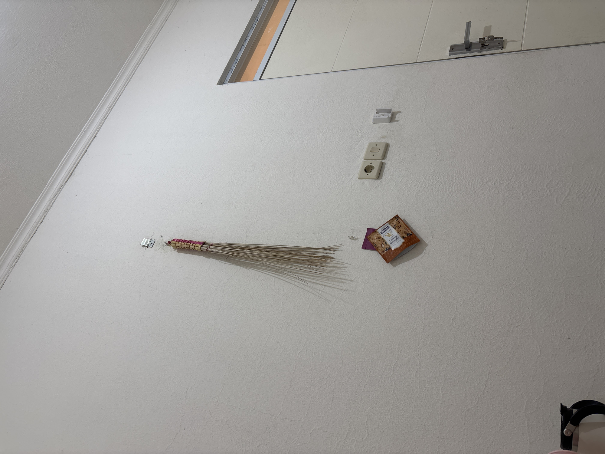 A small, dry-bristled broom is hanging from a metal hook attached to a textured white wall. Below the broom, a small, square package with a picture of dried leaves is also hanging from a hook. To the right of these items, there are two electrical outlets and a small white plastic holder on the wall. A portion of a white door with a silver handle and keys is visible on the right side of the frame. The ceiling molding is visible at the top of the image. The scene appears to be indoors, in a residential setting in Wanajaya, Indonesia.