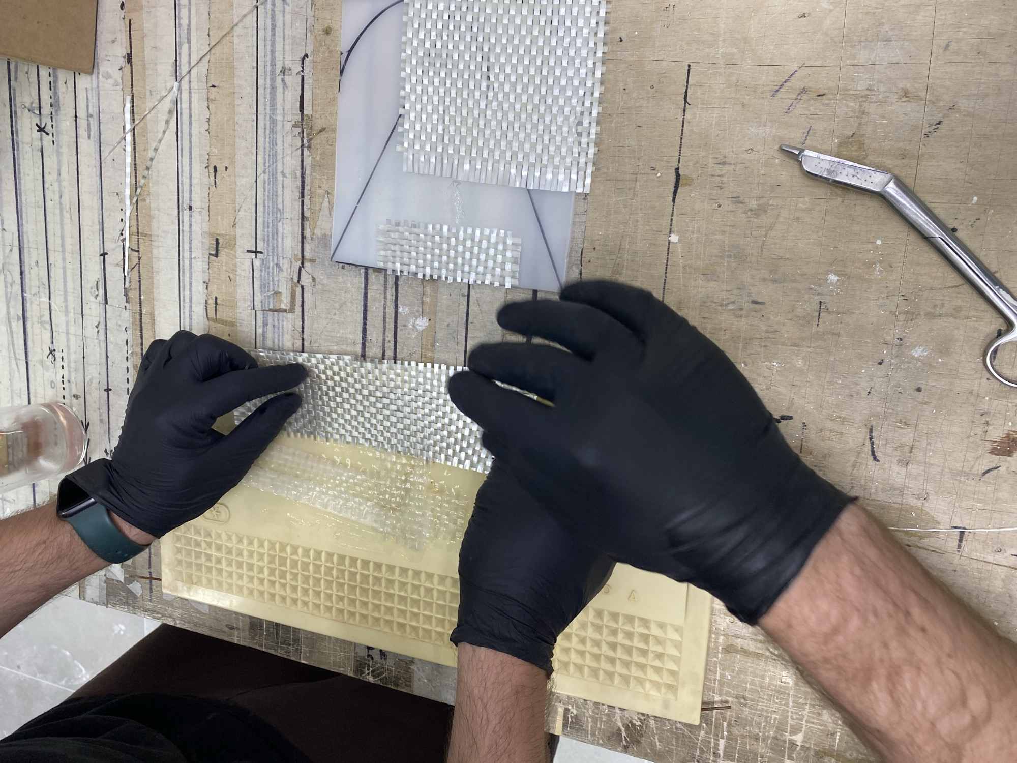 A top-down, medium shot captures two pairs of hands, each wearing black nitrile gloves, working on a wooden workbench covered with tape and markings. The hands are applying fiberglass fabric to a yellow textured mat and a clear plastic template. A glass container with clear liquid sits in the upper right corner. A pair of surgical scissors rests on the workbench in the lower right quadrant. The scene appears to be a workshop or crafting area. There is no discernible information in the image to suggest a specific location within the city of Vladimir, Russia.