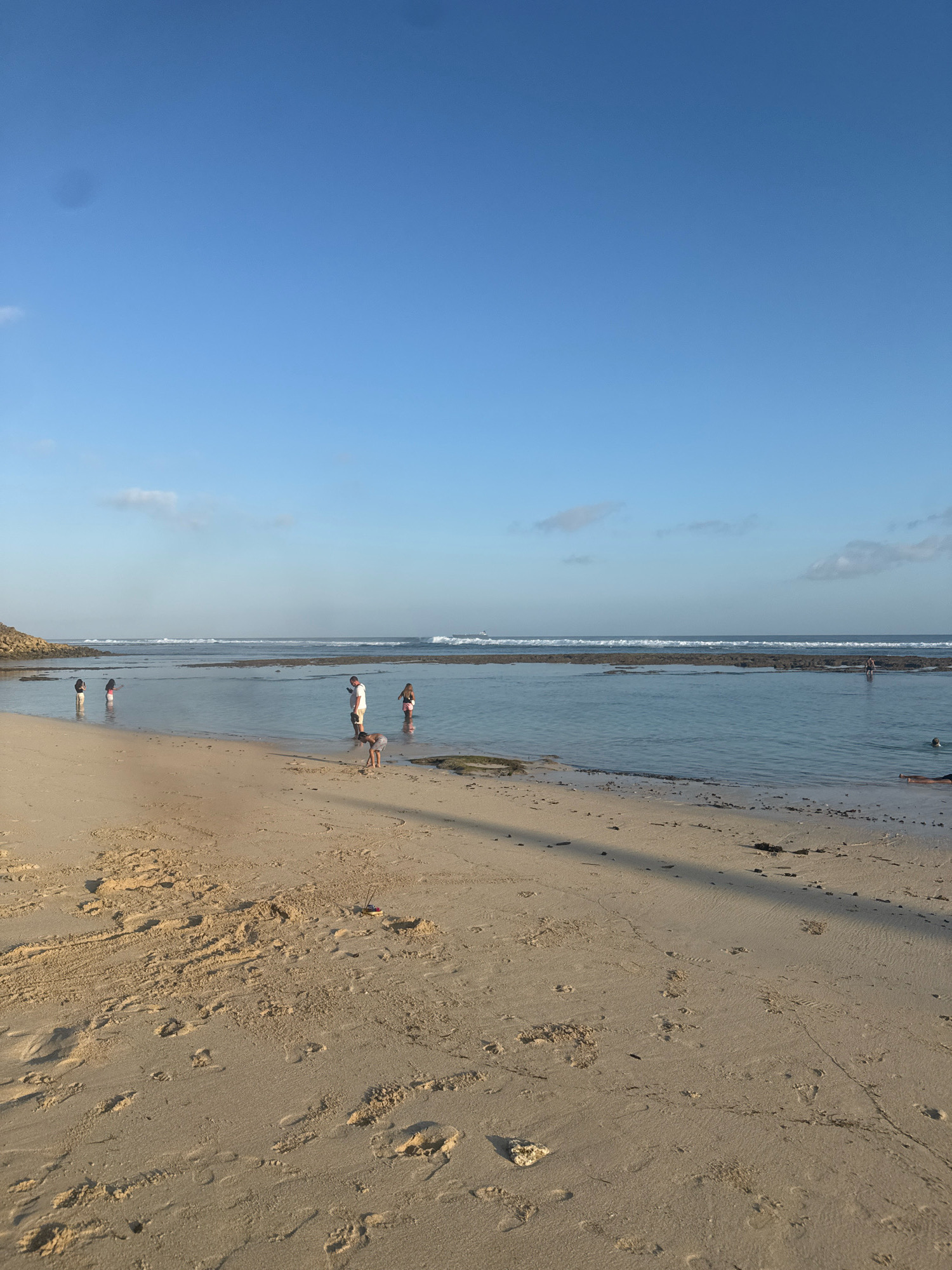 This image captures a serene beach scene in Kuta Selatan, Indonesia. The foreground is dominated by a sandy beach with numerous footprints etched into the surface, suggesting recent activity. A long shadow, cast by an unseen object, stretches across the sand. In the mid-ground, a shallow, calm body of water lies between the beach and the ocean. Several individuals are present in this area: a child is bent over, seemingly playing in the sand near the water's edge. Two adults, a man and a woman, stand in the shallow water. Further to the left, two other individuals, possibly children, stand in the water. In the distance, gentle waves break over a submerged reef, creating a white line against the blue ocean. The horizon is clear, meeting a vast expanse of bright blue sky with a few wispy clouds. A faint object, possibly a ship, is visible on the far horizon. The environment appears peaceful and idyllic, characteristic of a tropical beach setting.