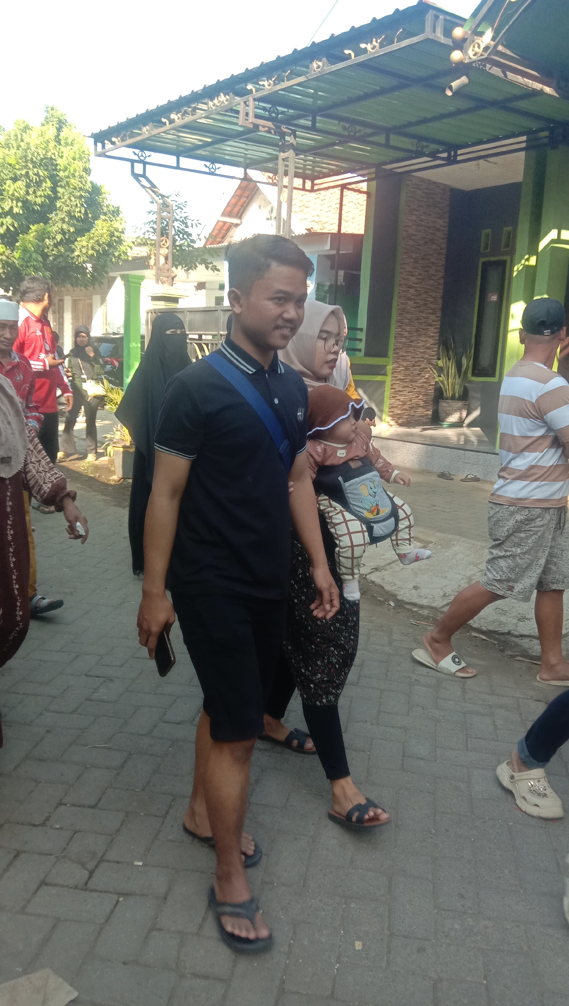 In Ngempit, Indonesia, a man in a black polo shirt and shorts walks towards the viewer, holding a dark smartphone. Beside him, a woman wearing a hijab and glasses carries a baby in a carrier on her chest. The baby is dressed in a brown onesie and hat. To the right, a man in a striped t-shirt and patterned shorts walks away from the viewer. Several other individuals are visible in the background, including a person in a red shirt and another wearing a black veil and robes. The scene takes place on a paved walkway, with buildings and trees lining the street. The architecture suggests a residential area, with a green tiled roof and a brown brick facade visible on one of the structures. The time of day appears to be daytime, with bright sunlight casting shadows on the ground.