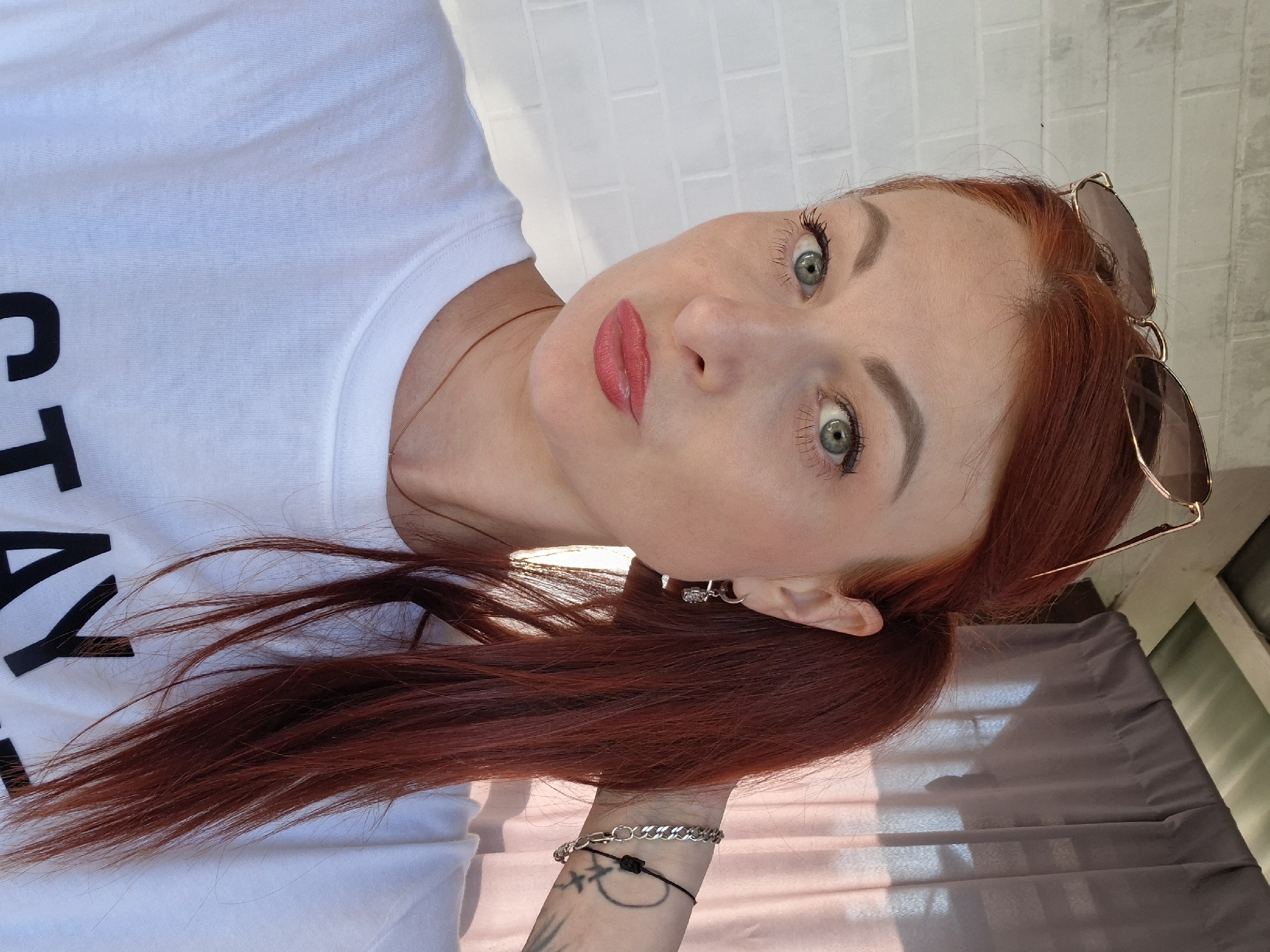 A woman with auburn hair pulled into a ponytail, wearing a white t-shirt and sunglasses pushed up on her head, poses for a selfie. She has green eyes, fair skin, and is wearing pink lipstick. She is also wearing a silver earring in her left ear and a silver chain bracelet on her right wrist. Behind her, a grey curtain hangs in front of a window. The image is taken outdoors against a white brick wall. The specific location in Myadzyel, Belarus is unknown.