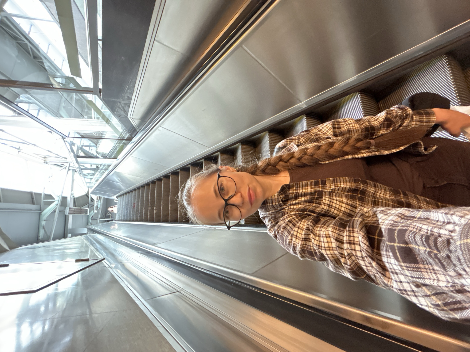A woman with glasses and a braid is riding an escalator. She is wearing a plaid shirt over a brown shirt. The escalator appears to be in a modern building with glass and metal accents. The scene is brightly lit, suggesting it is daytime. It is unclear if this location is in Saint Petersburg, Russia.