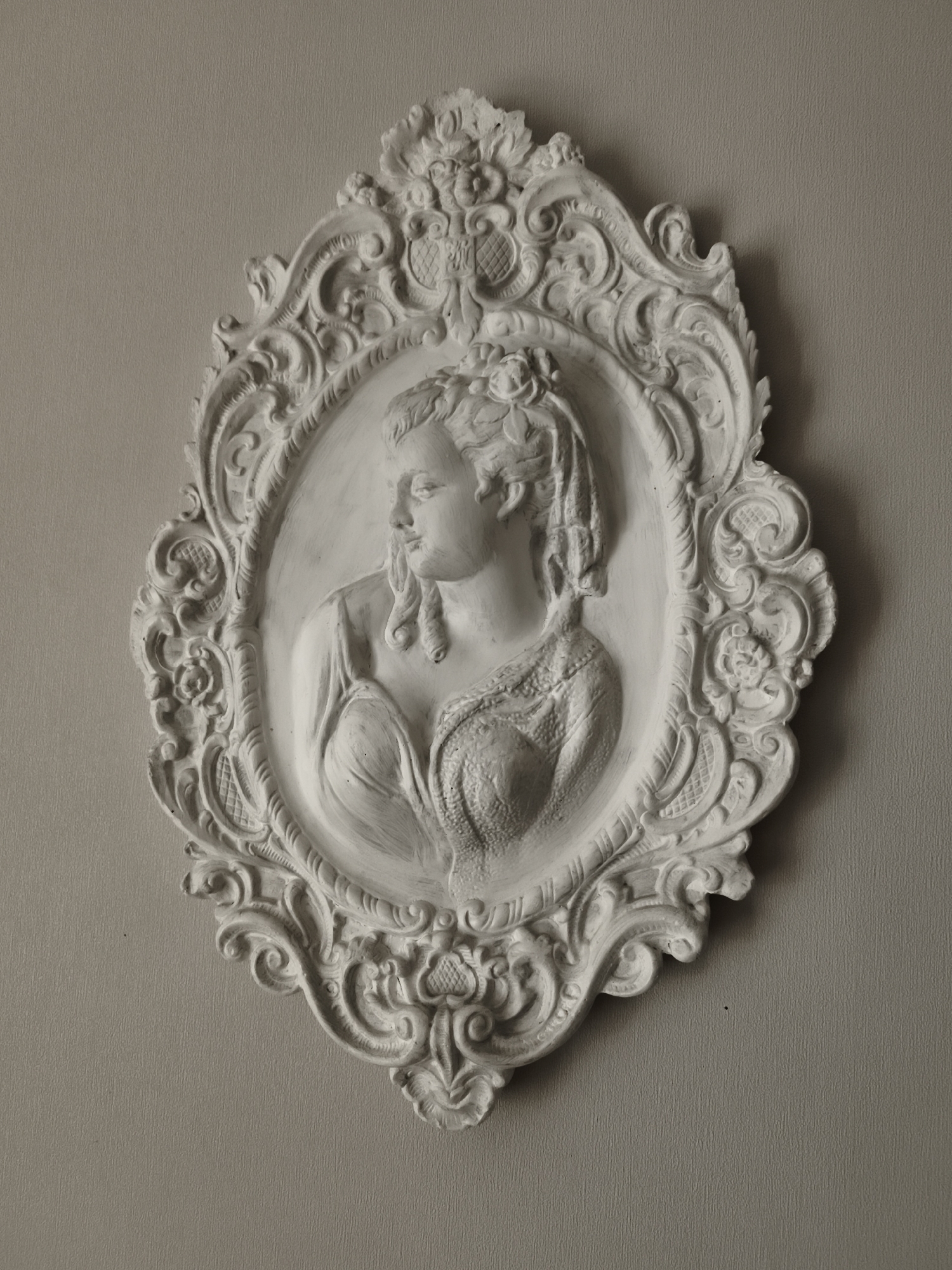 This is a studio photograph of a white plaster relief of a woman's bust, mounted on a plain, textured, light brown wall. The relief is oval-shaped and framed by an ornate, decorative border featuring scrollwork, floral motifs, and intricate detailing. The woman is depicted in profile, facing left, with her hair styled in curls and adorned with what appears to be a floral wreath. She is wearing a draped garment that reveals her shoulders and chest, with a detailed lace-like pattern across the upper bodice. The lighting suggests an indoor setting, with soft illumination highlighting the three-dimensional qualities of the relief and casting subtle shadows. The overall impression is one of classical or historical artistry, with the relief likely intended as wall decor. There is no visible text in the image.
