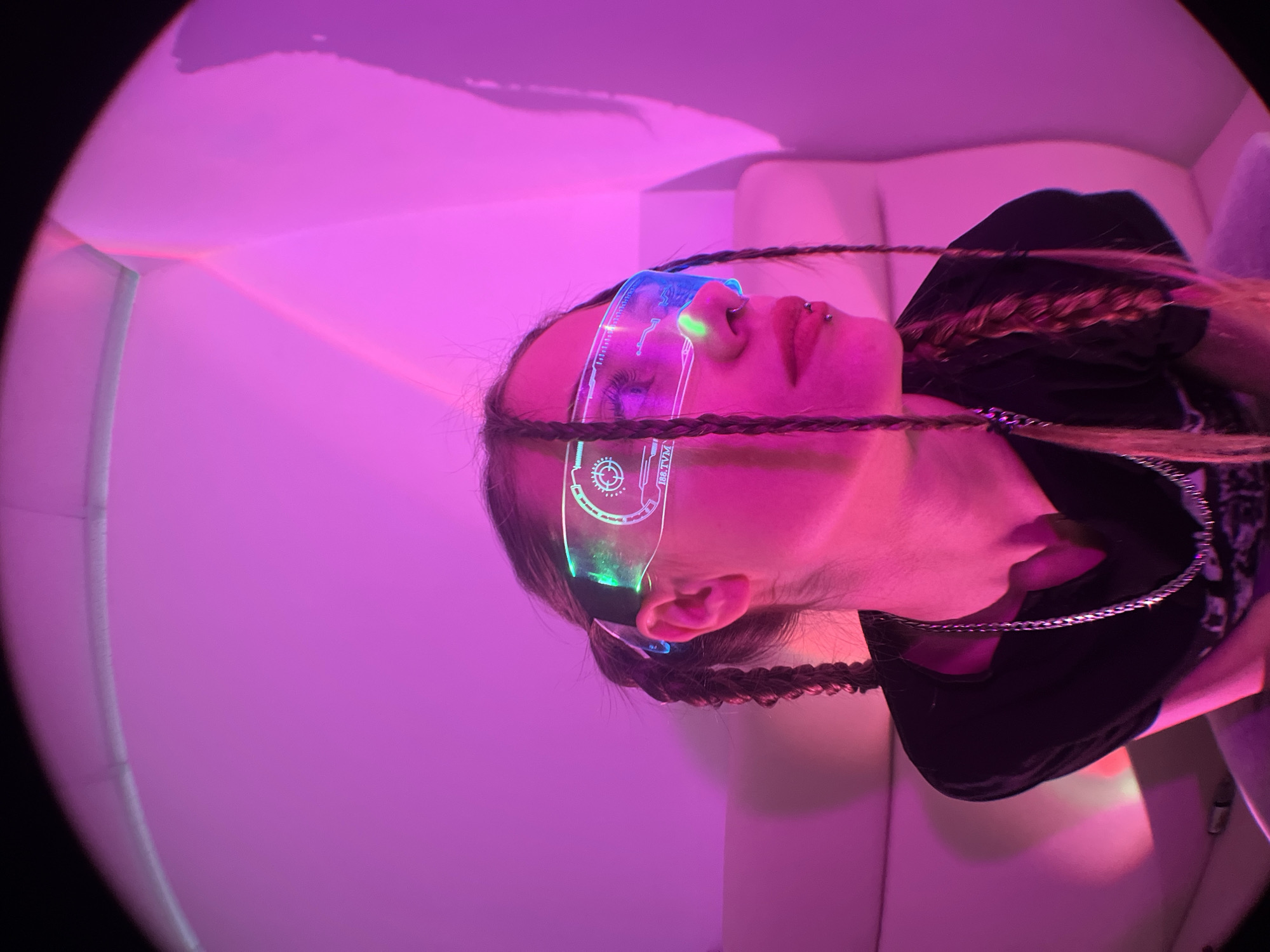 A person is shown from a low-angle perspective in a photograph that has been rotated counter-clockwise. The subject, who appears to be a young adult, is wearing futuristic-looking eyewear with illuminated designs. The hair is styled in braids and the person has piercings on their face. The subject is positioned against a backdrop of pink lighting that appears to emanate from the surrounding environment, possibly an enclosed space with white architectural elements. There is no indication that this photo was taken in or near Фоки, Russia.