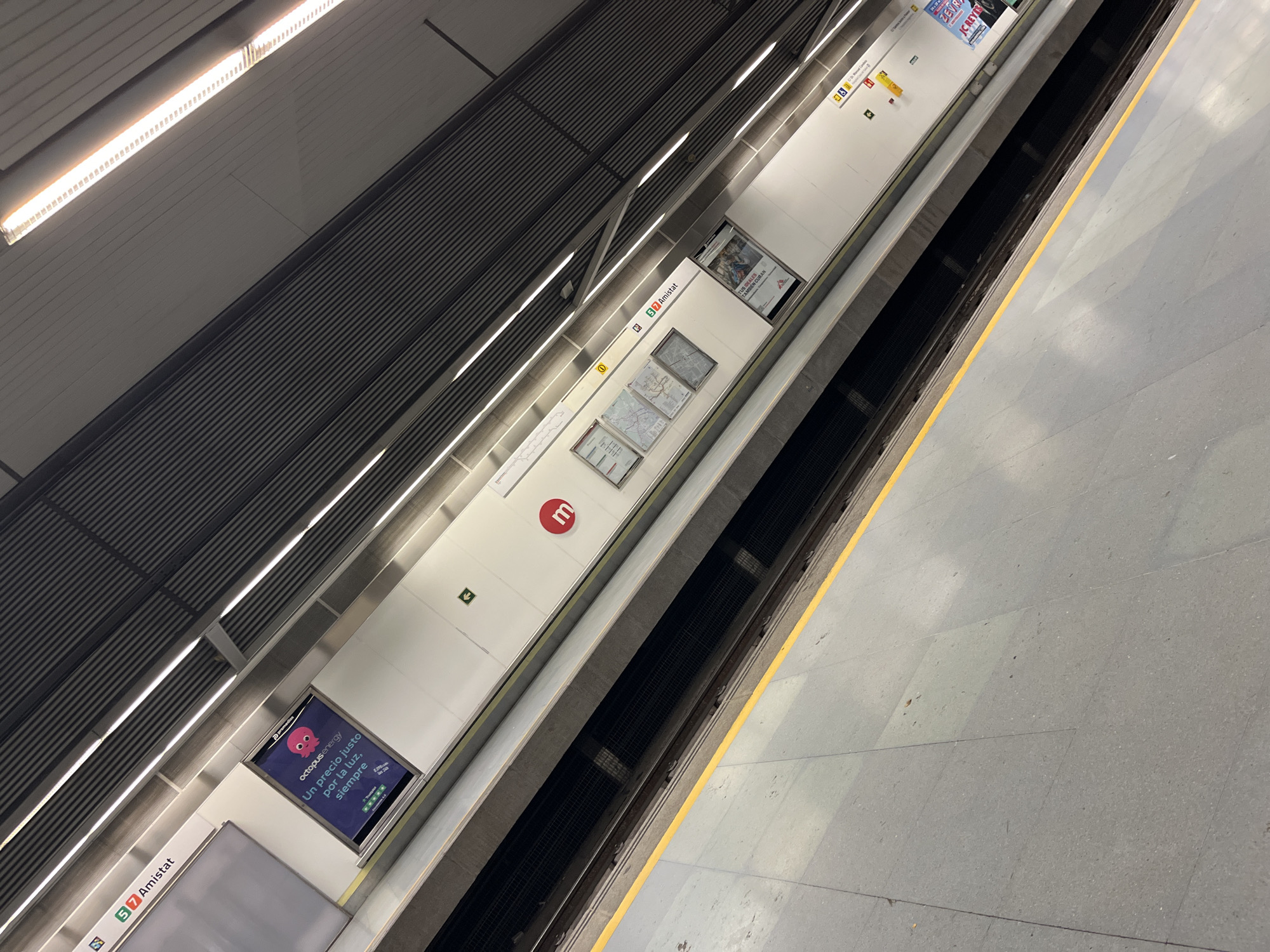 Here's a detailed description of the image:
**Scene:** The image shows an elevated, angled view of a metro station platform.
**Location:** The station is "Amistat" in the city of València, Spain. This is indicated by signage present in the image.
**Platform Details:**
* The platform is clean and has a modern aesthetic.
* There is a yellow safety line marking the edge of the platform.
* The station's symbol, a red circle with a white "m", is prominently displayed on the wall.
* Informational posters and advertisements are visible on the walls.
* One advertisement is for "Octopus Energy", and another one says "Tus ideales también curan"
* The lights consist of fluorescent tubes.
**Tracks:**
* The tracks are situated below the platform.
* It is not possible to see whether a train is present.
**General Environment:**
* The overall environment is well-lit.
* The station appears to be indoors.
* The platform surface is composed of gray tiles.