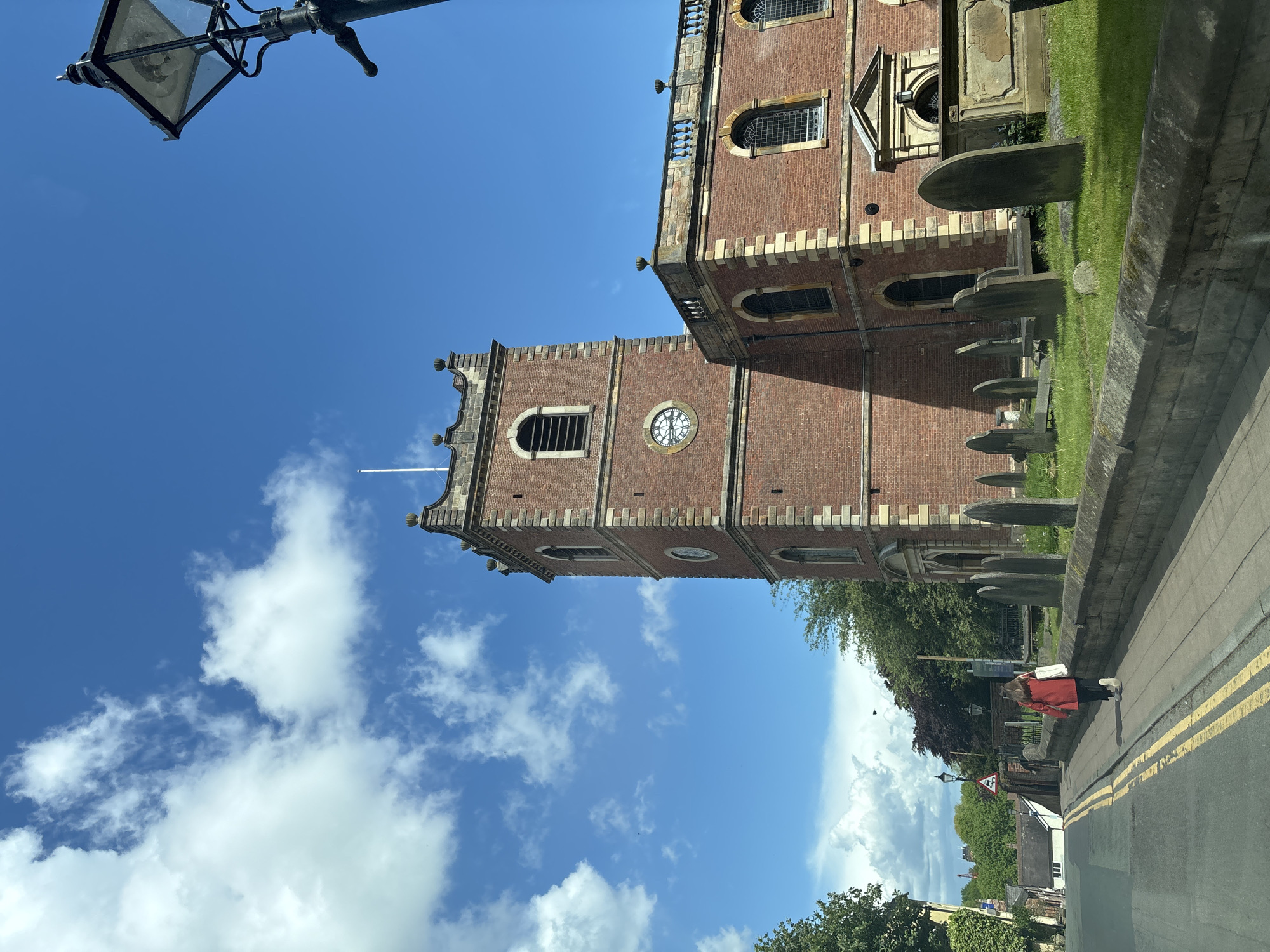 The image depicts St. John's Church in Knutsford, United Kingdom, from a low-angle street view. The red brick church tower with a clock face is visible against a bright blue sky with white clouds. A portion of the main church building is also visible, along with gravestones in the adjacent churchyard. A lamp post is in the upper left corner. A road with double yellow lines is in the foreground. A person in a red coat is walking on the pavement. Buildings and trees are visible in the background.