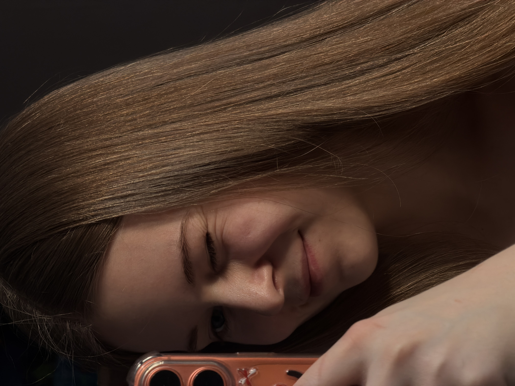 The image features a young woman lying down in an indoor setting in Perm, Russia. Her long, straight, light brown hair covers a significant portion of the frame, especially the upper and right sides. She is holding a smartphone, encased in a transparent orange or coral-colored case, with her left hand visible in the lower right corner. The phone's camera lenses are clearly visible on the case.
The woman has fair skin and a playful, amused expression. Her lips are curved into a slight smile, and her right eye is partially closed, creating a winking or squinting effect. Her left eye is visible, looking towards the phone. The lighting is dim, suggesting an evening or nighttime scene or a dimly lit room, with a dark, unidentifiable background that enhances focus on the subject. The scene conveys a relaxed and personal moment, possibly of her engaging with her phone, perhaps taking a selfie or video. No specific location cues other than the indoor setting are discernible. There is no visible text in the image.