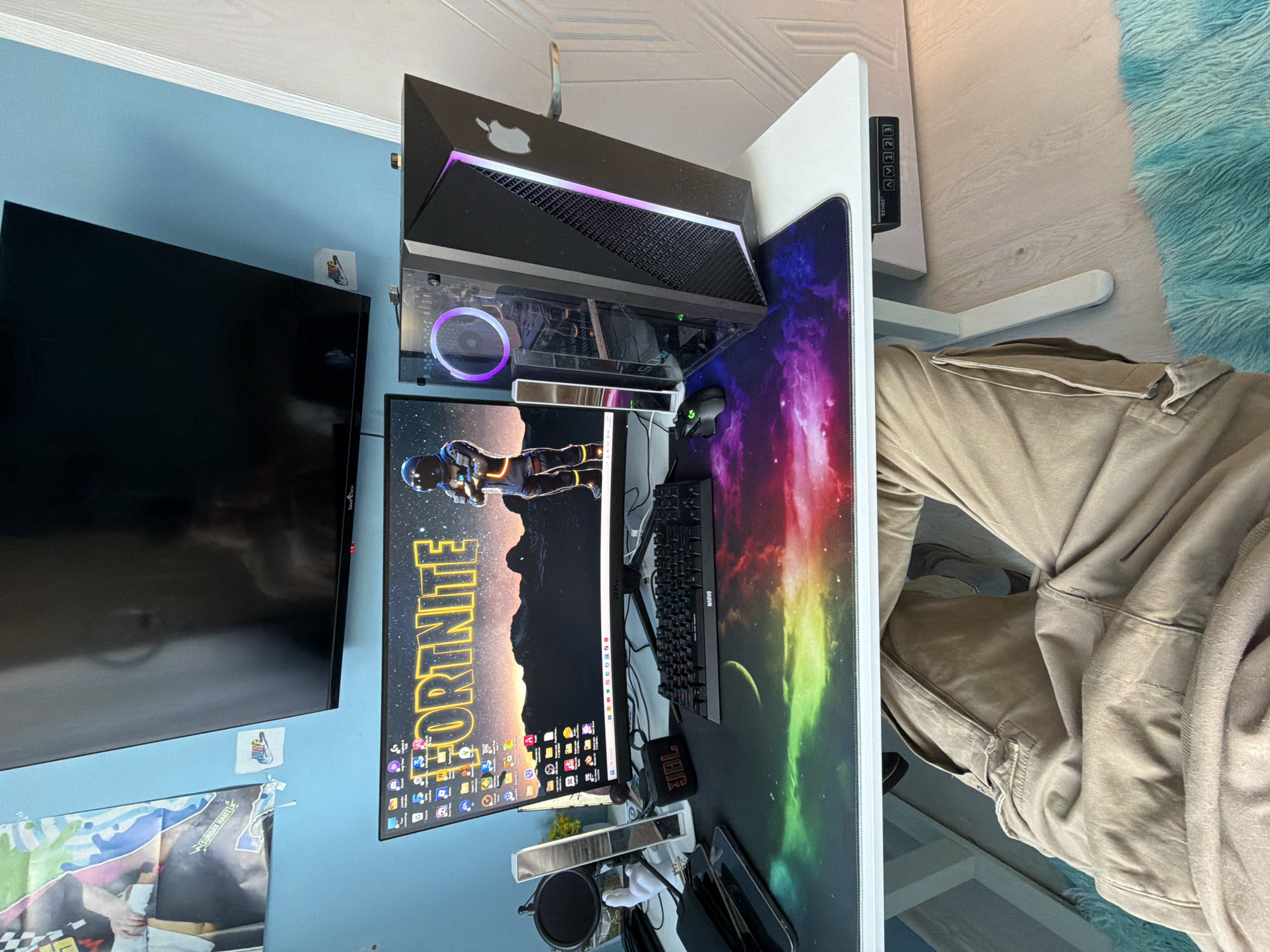 The image shows a desk setup against a light blue wall. A black flat-screen TV hangs on the wall, along with a poster featuring a rainbow-colored image. Below the TV is a monitor displaying a Fortnite-themed wallpaper, positioned vertically. A black keyboard and mouse are on a large, colorful mousepad. A black computer tower with a purple light strip and a visible cooling fan sits on the desk. A white desk frame supports the desk surface. A microphone with a pop filter is also present. Part of a person's body, wearing tan pants, is visible to the right side of the image. The location is unknown.