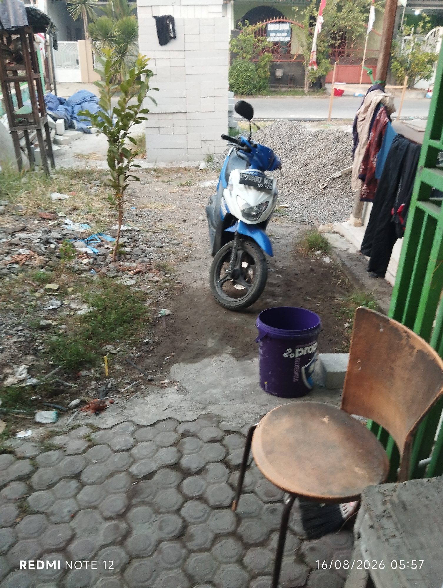 The image depicts an outdoor scene in what appears to be a residential area in Gempol, Indonesia. In the foreground, a hexagonal paver walkway leads to a rustic wooden chair with a round seat. To the left of the chair is a purple bucket. Behind the chair and bucket, a blue and white scooter is parked on a dirt and gravel surface. The scooter has a license plate that reads "L 2800 WL". Further back, a small tree grows near a wall constructed of white cinder blocks. Beyond the wall, a street is visible with houses and vegetation. The background includes a glimpse of a fence and some laundry hanging on a line. The overall environment suggests a casual, possibly unfinished, outdoor space.