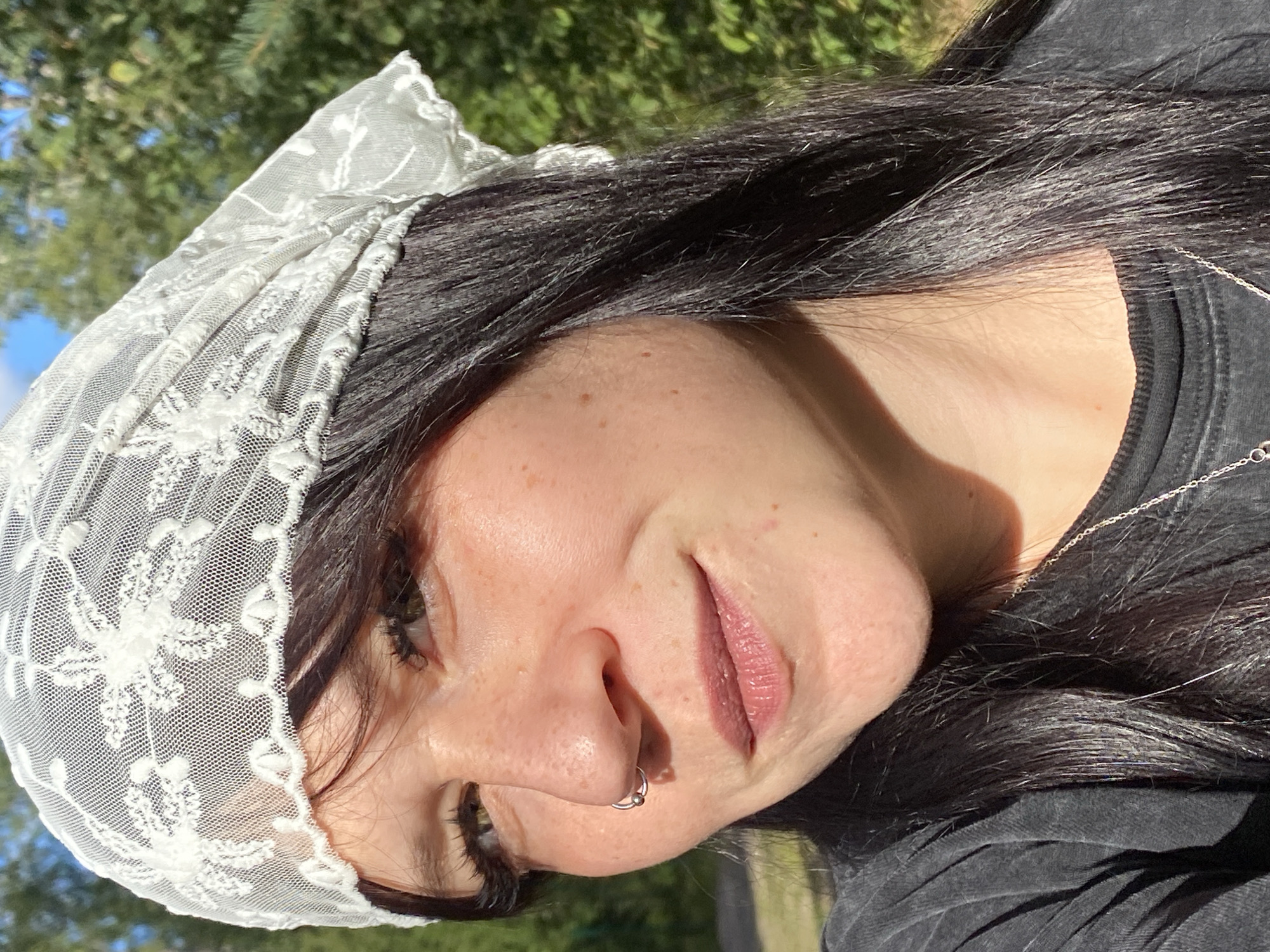 In the city of Krasnoflotskoe, Russia, a close-up, profile shot captures a young woman with dark hair and fair skin. She is wearing a white lace headscarf and a dark t-shirt, with a silver necklace visible. The woman has a nose ring and her eyes are looking to the left. She has freckles scattered across her cheeks and nose, and her lips are painted a subtle pink. Sunlight illuminates her face, creating shadows and highlighting the textures of her skin and the lace. Behind her, out-of-focus greenery suggests an outdoor setting, possibly a park or a yard, with a clear blue sky visible in the upper portion of the frame.