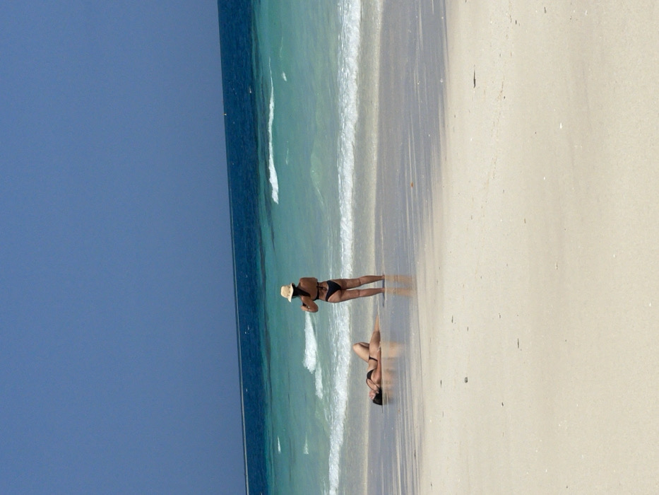 A sun-drenched beach scene features azure skies and turquoise waters meeting a sandy shoreline. Two women are present: one standing near the water’s edge, wearing a dark swimsuit and a straw hat, and another lying prone on the beach. The reflection of the standing woman appears in the wet sand, creating a double image. The scene exudes a tranquil and sunny vacation atmosphere.