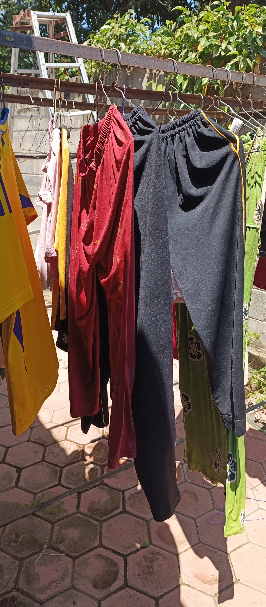 In an outdoor setting, likely a backyard or courtyard, clothing hangs on a metal drying rack. The clothes are suspended by hangers, including a bright yellow t-shirt with blue stripes, a light pink garment, a yellow garment, a dark red pair of pants with a subtle pattern, a pair of black pants with a yellow stripe along the side, and a green patterned garment. The ground below is paved with hexagonal tiles. In the background, a ladder leans against a wall, and lush green foliage is visible against a bright blue sky. The scene is illuminated by bright sunlight, casting distinct shadows of the clothing onto the tiled ground.