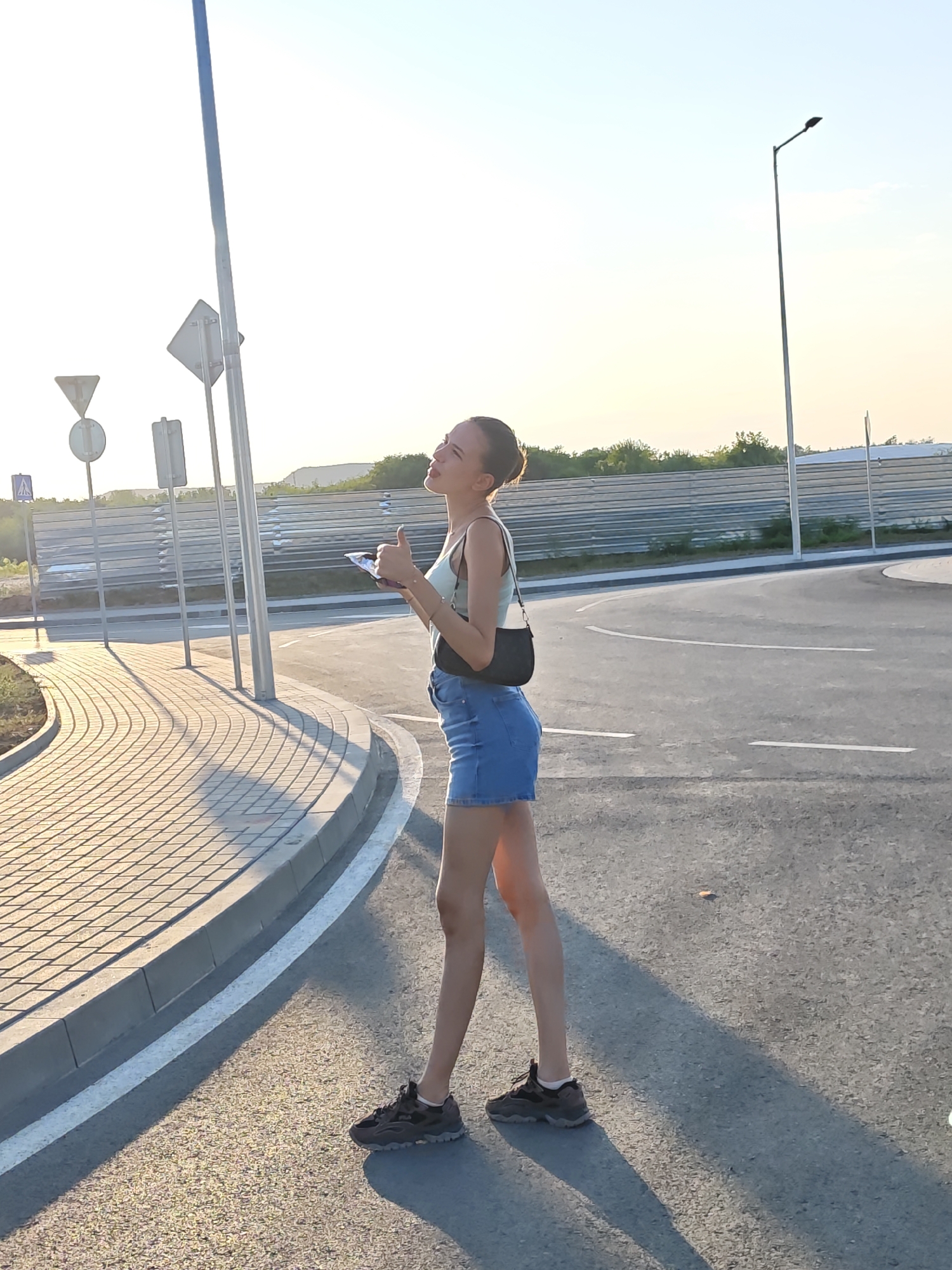 A young woman stands on an asphalt road, her body angled slightly away from the camera, with her head turned upwards and to the right. She is wearing a light blue tank top, a denim skirt, and dark athletic shoes. A dark purse is slung across her body. Her right hand is raised, with her thumb up, and she holds a small object in her left hand. The woman is positioned on the right side of the image, with a paved walkway and curb to her left. In the background, a metal fence and some trees are visible, under a bright, hazy sky. Streetlights and traffic signs are also present in the scene. The overall lighting suggests it is either early morning or late afternoon. The specific location in Lovech, Bulgaria, cannot be determined from the visual information alone.