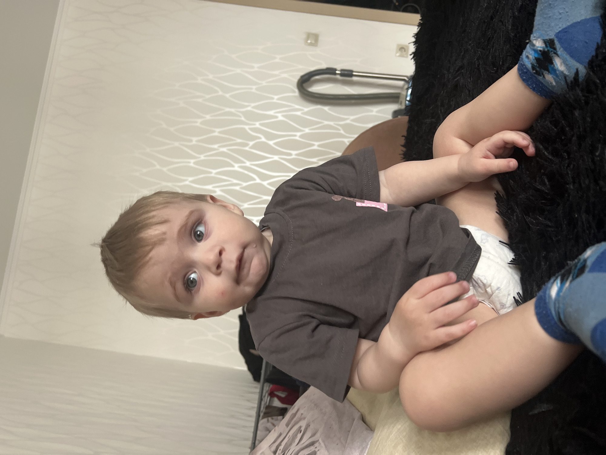An indoor photograph, taken in Perm, Russia, features a young toddler with light blonde hair and striking light blue eyes, looking directly at the camera with a wide-eyed, curious, and slightly puckered expression. The child is wearing a dark grey t-shirt with a small, indistinct pink design on the left chest area and a white disposable diaper. They are seated on a soft, black, shaggy surface, possibly a blanket or pillow. Part of a leg wearing a blue sock with a diamond or argyle pattern is visible on the right.
The background shows a light-colored wall with a subtle, wavy patterned texture. A vacuum cleaner hose and nozzle are visible leaning against the wall in the upper mid-ground, along with electrical outlets. The setting appears to be a domestic interior, likely a living room or bedroom, under even indoor lighting conditions. The image captures a moment of direct interaction between the child and the photographer.