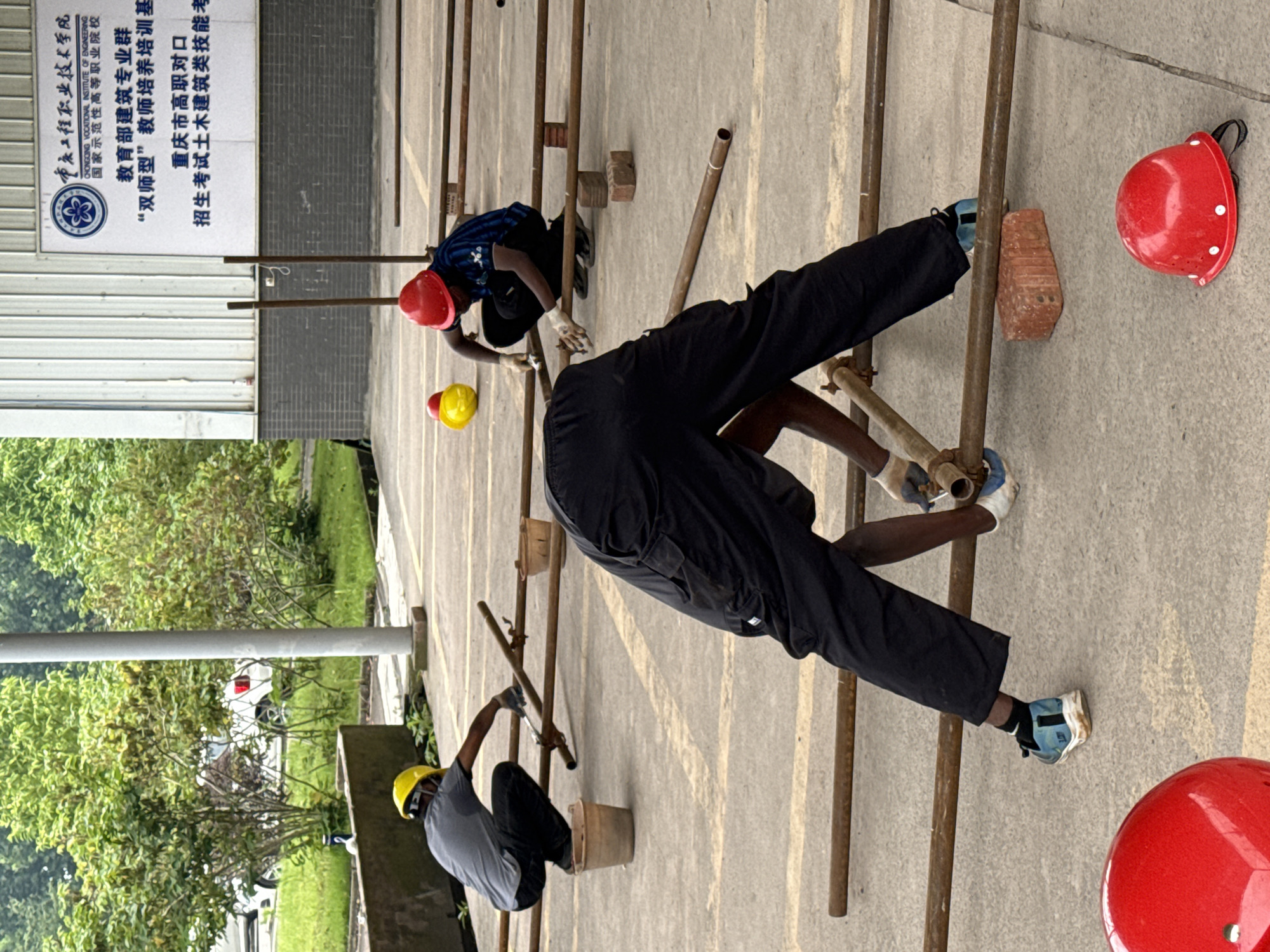 In Jiangjin, China, three individuals are engaged in a construction-related training activity outdoors on a paved surface. In the foreground, a person wearing black pants and white gloves is bending over, working with metal pipes. Their footwear consists of blue and white athletic shoes. To the left of this individual, a person in a yellow hard hat and grey shirt crouches down, also interacting with pipes and holding a tool. They wear dark pants and are positioned near a brown bucket. Further to the right, another person in a red hard hat and a blue and black striped shirt kneels, manipulating metal pipes. Yellow and red hard hats are visible on the ground nearby. The background features a building with signage and greenery, including trees. The overall environment suggests an educational or training facility, possibly associated with vocational engineering or construction, given the presence of safety gear and construction materials.
