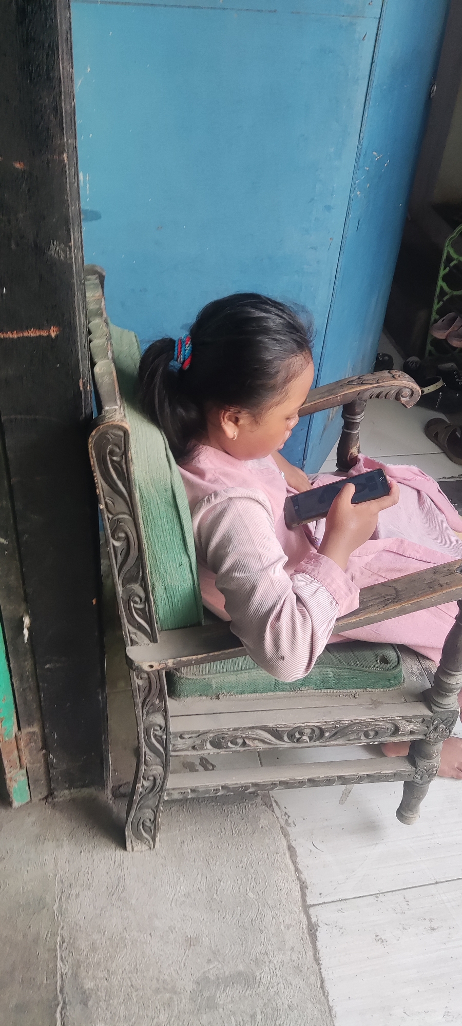 A young girl, with dark hair pulled into a ponytail, sits on a wooden chair with faded green upholstery. She wears a pink long-sleeved shirt. She is holding a black smartphone in her right hand and appears to be looking at it. The chair is ornate with carved details and is placed on a concrete floor. In the background, a blue wall is visible, and to the left, a dark, wooden structure. The setting appears to be an outdoor or semi-outdoor area, possibly in a residential area in Gebang, Indonesia, given the style of the chair and the rustic environment.