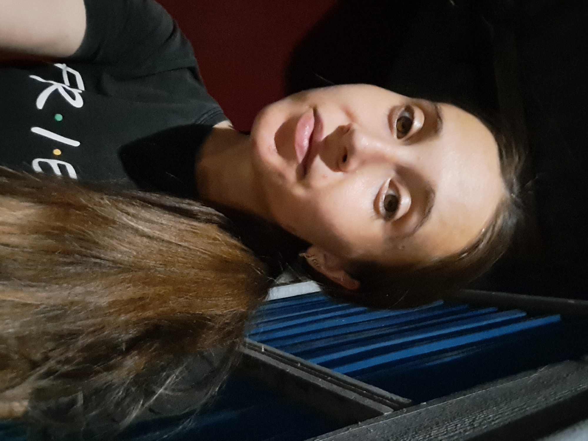 A young woman with light brown hair tied in a ponytail is looking towards the camera. She is wearing a black t-shirt with the word "FRIENDS" printed in white, with colored dots between the letters. The background is dark, with some vertical blue and gray structures on the right side and a dark red wall on the left. The image appears to be a selfie taken in Vypasne, Ukraine.