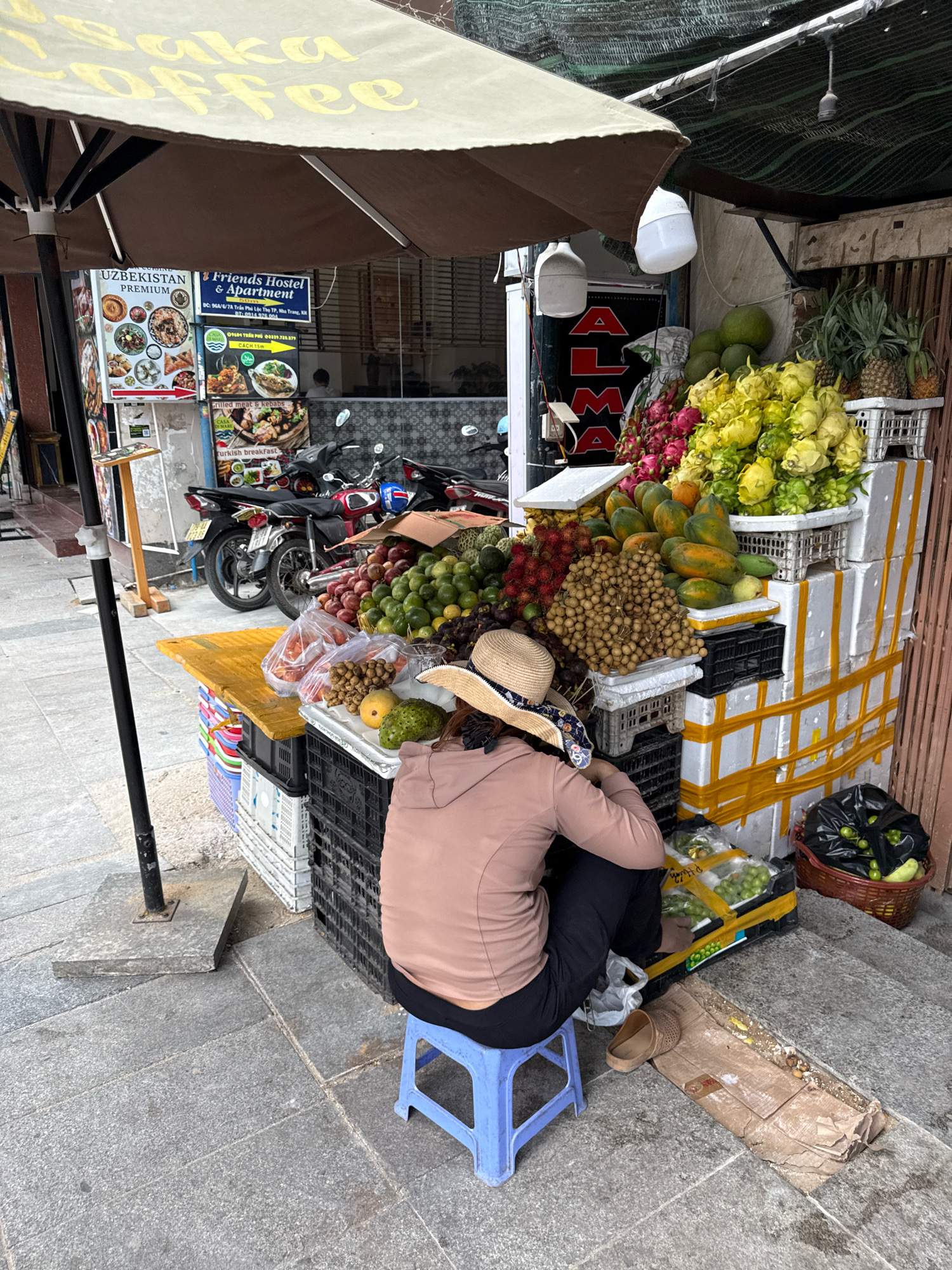 An outdoor fruit stand is set up on a sidewalk in Nha Trang, Vietnam, under a large brown umbrella with a yellow sign that reads "Coffee". A woman wearing a straw hat and a light brown hoodie sits on a blue plastic stool, facing away from the camera, tending to the fruit. The stand displays a variety of fresh fruits, including pineapples, dragon fruit, rambutans, longans, papayas, and green grapes, arranged on crates and styrofoam boxes secured with yellow tape. Behind the fruit stand, several motorcycles are parked on the street. A sign on the building behind the stand advertises "Friends Hostel & Apartment". The sidewalk is paved with gray tiles. The environment appears to be a typical street scene in a commercial area.