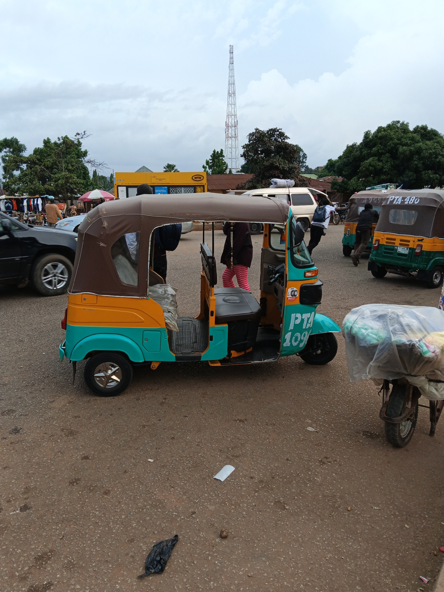 The image captures a street scene in Bukuru, Nigeria, likely a marketplace or transportation hub. The foreground is dominated by a colorful auto-rickshaw, painted in teal and yellow with a brown canopy. It has "PTA 109" written in large white lettering on its side. Behind it, another auto-rickshaw, this one green and yellow, is visible with "PTA-480" on its rear. A third auto-rickshaw, mostly obscured by the others, is to the left.
Several people are present, some appearing to interact with the auto-rickshaws, possibly as drivers or passengers. One person in the teal and yellow auto-rickshaw seems to be interacting with someone outside the vehicle. Another individual in a white shirt is leaning into the auto-rickshaw on the right. A person wearing a dark jacket is partially visible behind the green and yellow auto-rickshaw. Further in the background, there are market stalls with clothing displayed, suggesting a bustling commercial area.
A yellow building with an MTN logo is visible behind the market stalls, indicating the presence of a telecommunications company. A tall, lattice tower, likely a radio or cellular tower, stands prominently in the sky, which is overcast with clouds. The ground is paved with rough asphalt or gravel. Scattered on the ground are a black plastic bag and some white debris. The overall impression is one of everyday life and commerce in a Nigerian city.