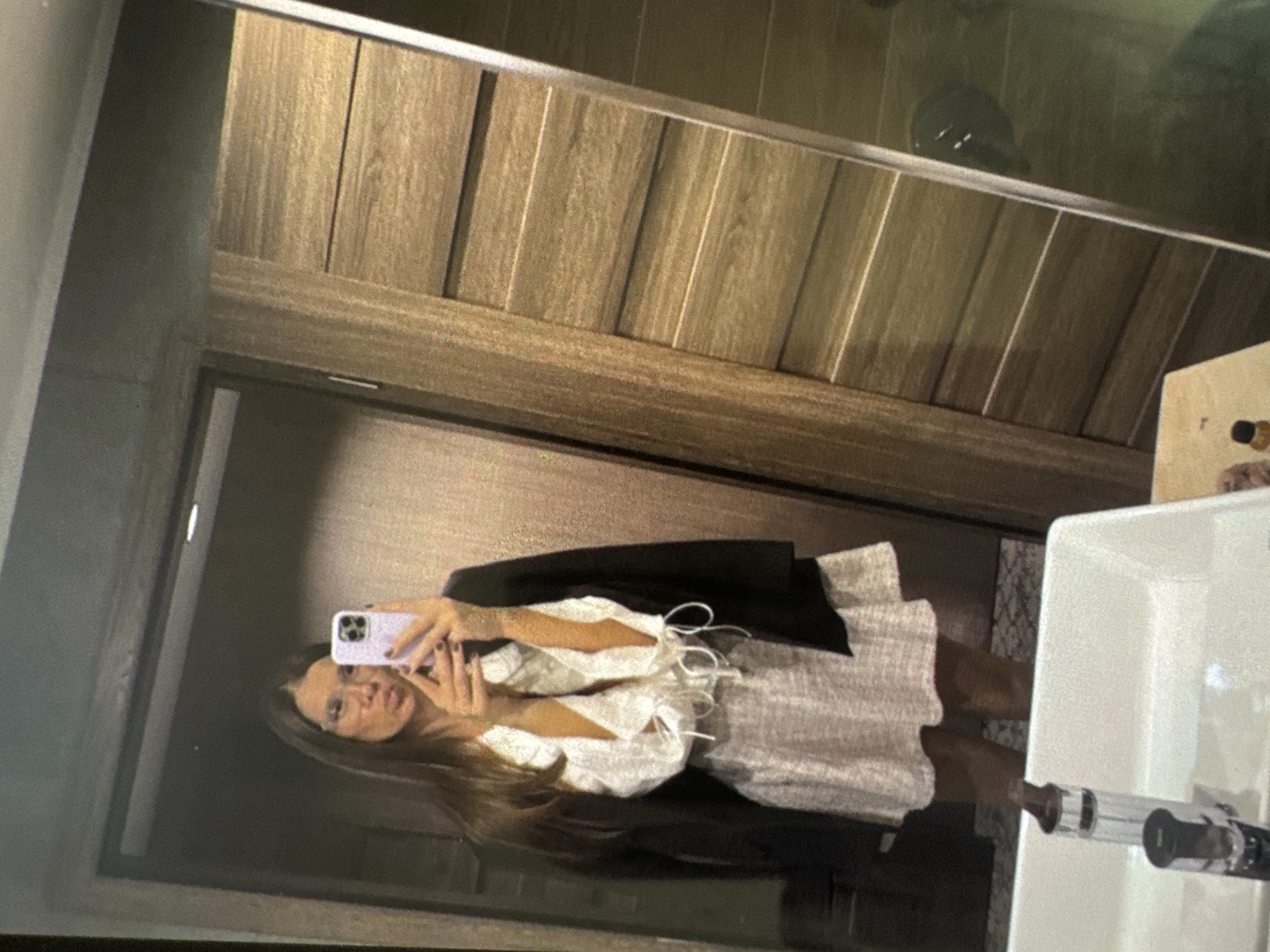 In Vratsa, Bulgaria, a female individual is captured in a mirror selfie within an indoor setting. The subject, with long brown hair, wears clear-rimmed glasses and has dark nail polish. Her lips are pursed. She is dressed in a white, long-sleeved top featuring ruffled details and white ties on the cuffs. A black blazer is draped over her shoulders, and she wears a light-colored, possibly patterned, mini skirt. She holds a light purple smartphone with a visible camera array, likely an iPhone, in front of her to capture her reflection.
In the foreground, a white rectangular sink with a dark, modern faucet is visible. An elongated metallic object, consistent with an electronic cigarette or vape pen, stands upright within or on the edge of the sink. Small bottles are positioned on the counter surface adjacent to the sink.
The background, as reflected in the mirror, features architectural elements. To the right of the subject, a wall or door segment is paneled with horizontal wooden slats. A thin, metallic vertical strip borders this segment. To the left, a darker, possibly gray or black doorframe or wall is reflected. The ceiling, visible at the top, is dark and includes a single recessed light fixture. The lower right portion of the reflection shows a dark, possibly stone or tiled, floor.