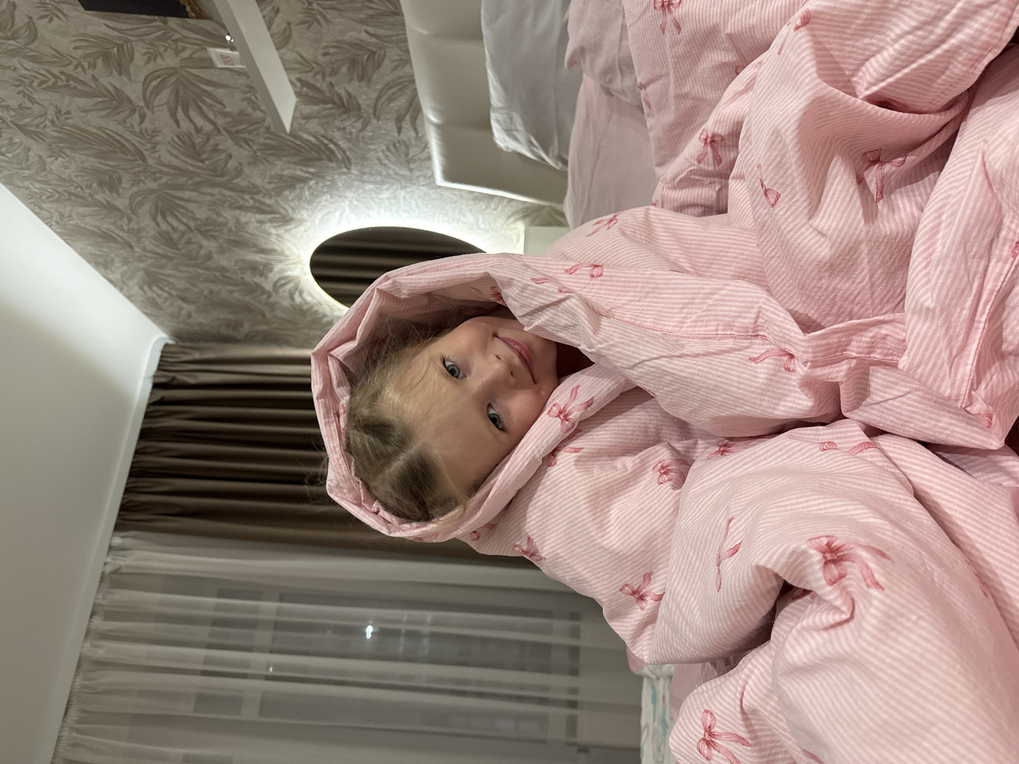 A young girl with fair skin and blonde hair is nestled in a pink and white striped blanket adorned with small bows. She is looking towards the camera with a slight smile. The setting appears to be a bedroom. The room features a patterned wallpaper, a round illuminated mirror, and sheer curtains over a window. The image was not taken in Izhevsk, Russia.