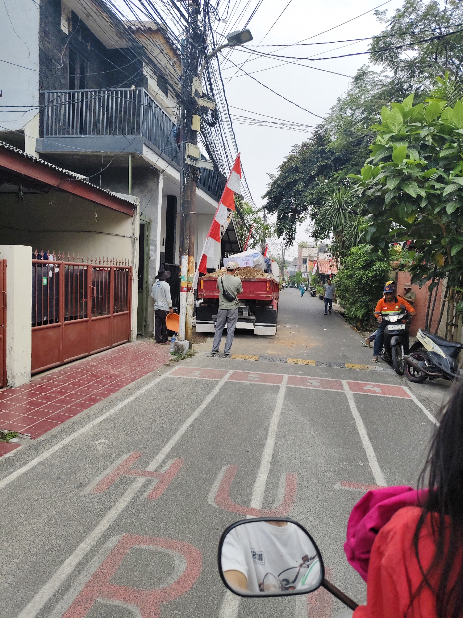 The image depicts a street scene in South Jakarta, Indonesia, with a red and white Indonesian flag visible on a pole. The street is paved and marked with red and white lines, possibly indicating lanes or a game area. In the foreground, a person is riding a motorcycle, reflected in the rearview mirror, wearing a white t-shirt and a pink garment. A red truck, loaded with soil or construction material, is parked on the right side of the street, with two men standing near it. One man, wearing a hat and a light-colored shirt, holds an orange shovel. Another man, wearing a green shirt and a backpack, is also present. A black motorcycle is parked behind the truck. The street is lined with buildings on the left and trees and more buildings on the right. Overhead, a dense network of electrical wires is prominent. A person in an orange shirt is seated on a motorcycle further down the street.