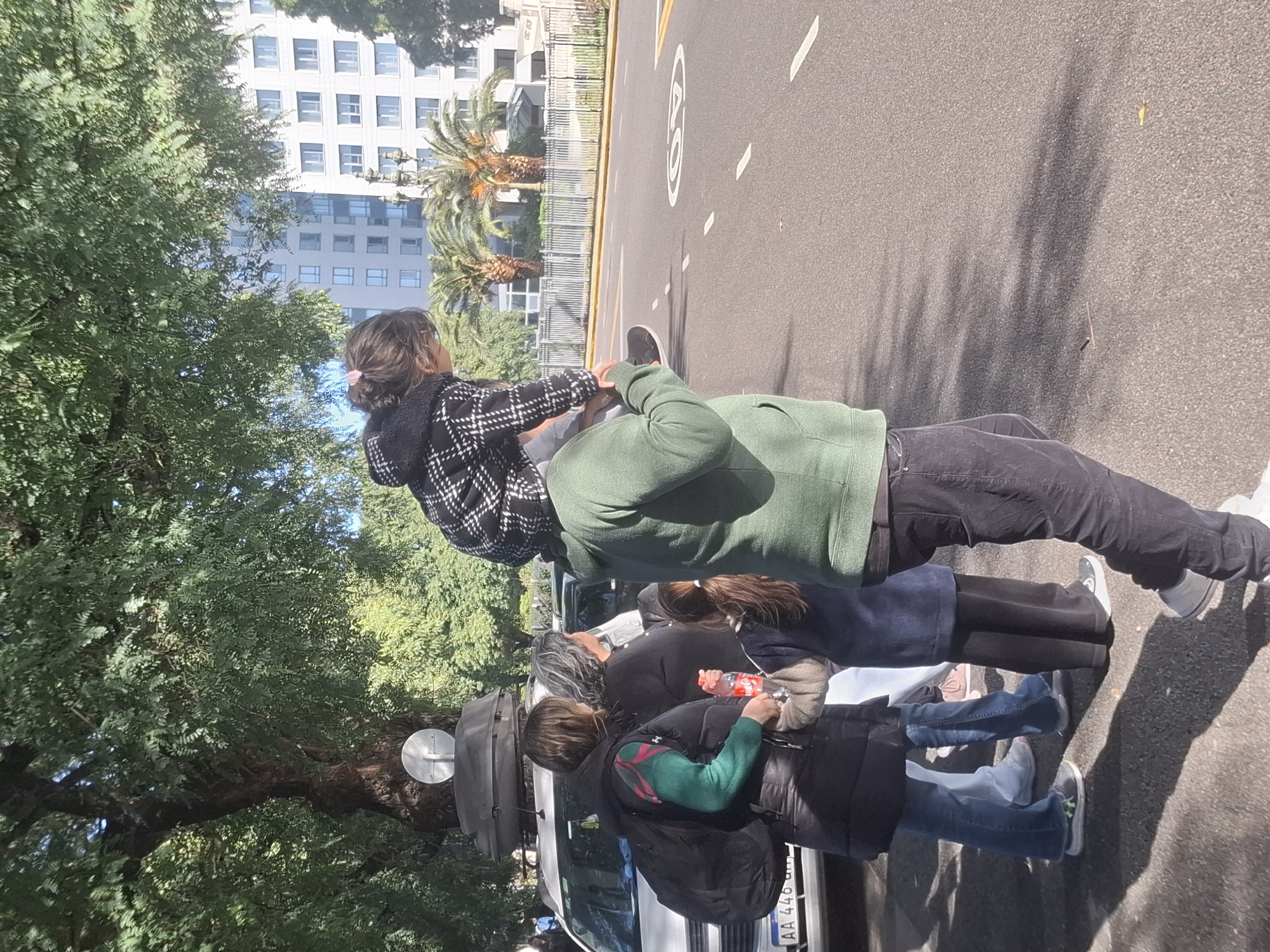 The scene captures a group of people, presumably a family, near a street in Buenos Aires, Argentina. A child is being held by an adult, while other adults stand nearby. There is a vehicle visible, potentially a van or small truck, parked adjacent to the sidewalk. The environment includes trees, a building in the background, and street markings on the asphalt. The individuals are dressed in casual attire, suggesting a relaxed, everyday setting.