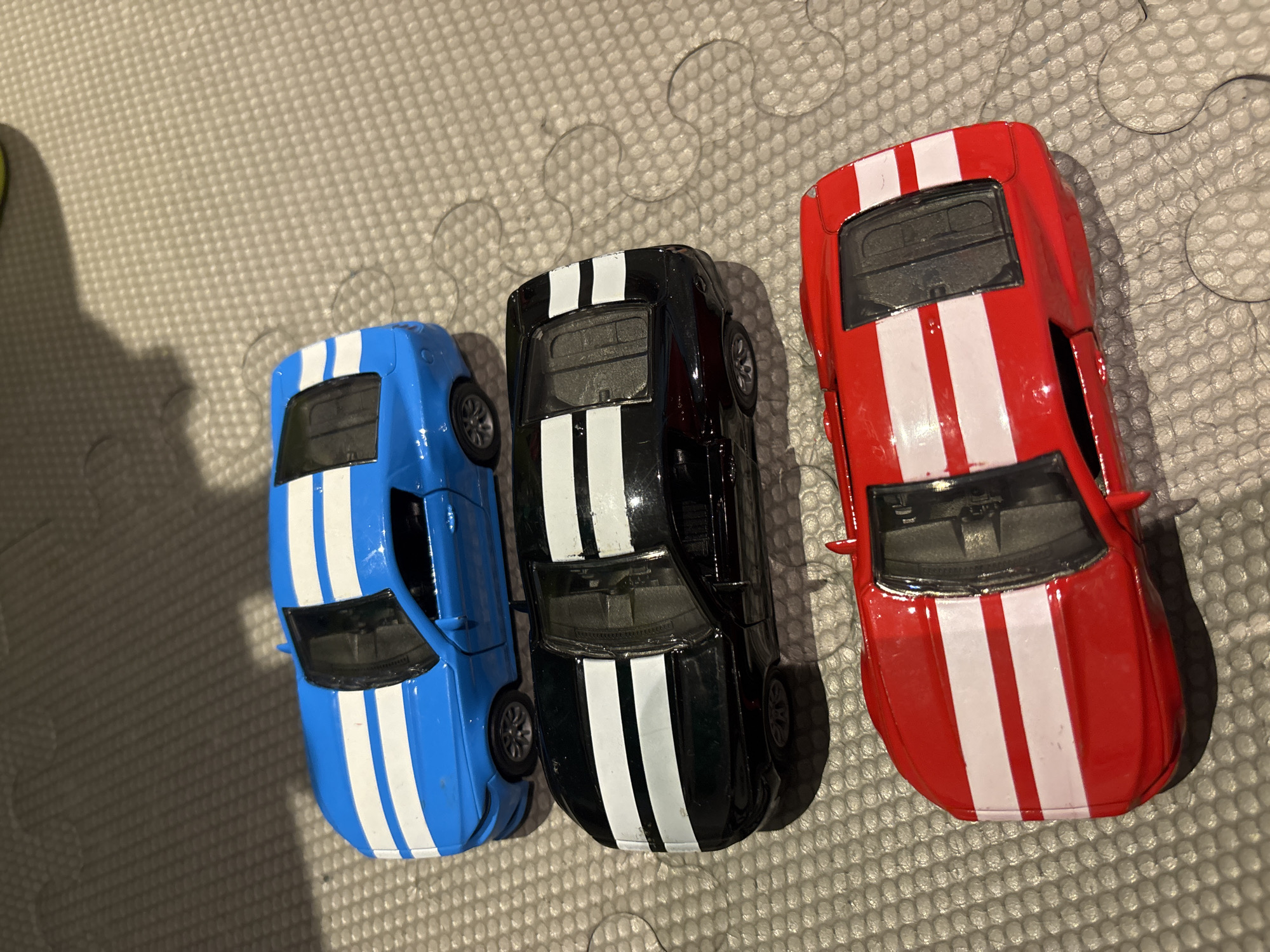 The image shows three toy cars, a blue, a black, and a red one, arranged in a stack. Each car has two white racing stripes on its hood and roof. They are placed on a grey, textured surface, possibly a play mat or a textured floor. The lighting suggests it is indoors. There is no indication of the location being Dubai, United Arab Emirates, or any specific city. There are no people or actions depicted in the image.