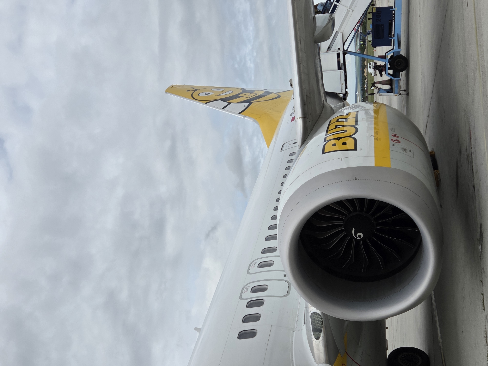 The image shows the side of a Buzz airline airplane parked on the tarmac. The airplane is white with yellow accents and features the Buzz logo on its tail, which depicts a smiling bee. The prominent engine of the airplane is visible in the foreground, with its fan blades clearly discernible. A set of stairs is attached to the aircraft, and a few people are visible near the top of the stairs, appearing to board or disembark. In the background, airport equipment, including a blue cart with the number "200" visible, and a white van are present. The scene is set against a cloudy sky. The image was taken in Aksakovo, Bulgaria.