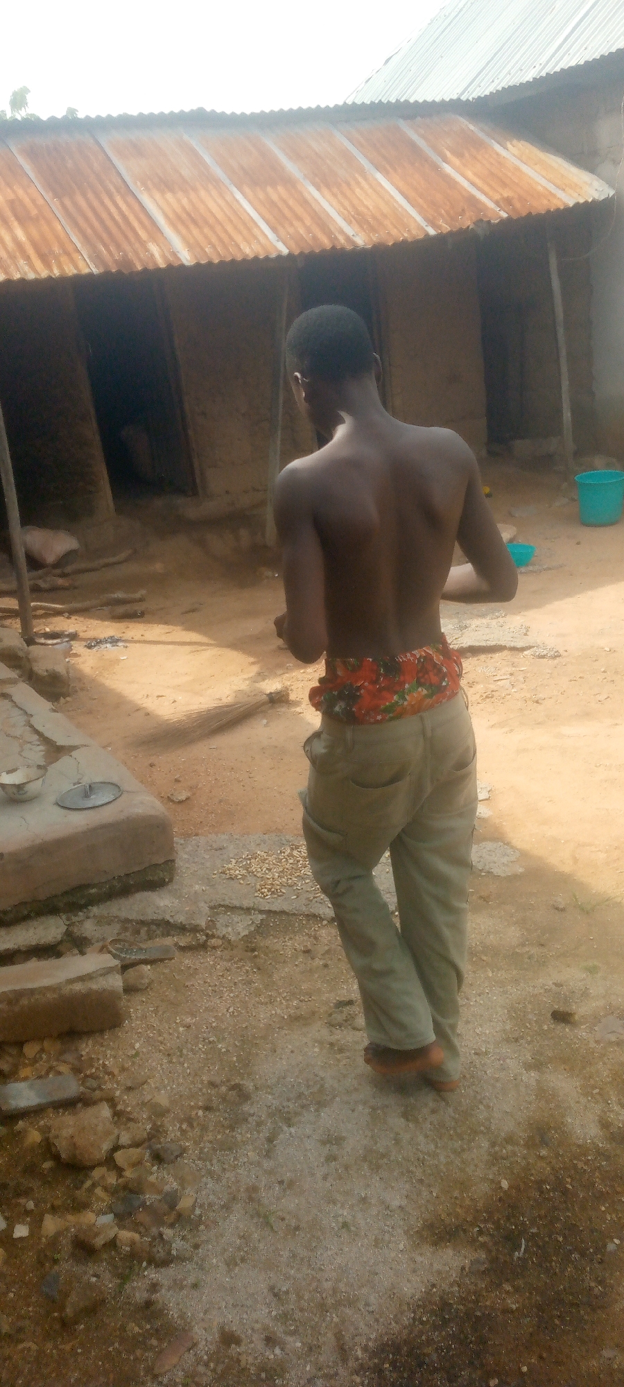 A young Black man is pictured from behind, shirtless and wearing tan pants with a brightly patterned waistband. He stands in a dirt yard in front of a rustic, dilapidated building with a corrugated metal roof. The ground is covered in sand, pebbles, and scattered debris. To the left, a concrete structure holds a small metal bowl and lid. A broom leans against the building to the right of the man. In the background, another building with a tin roof is visible, and a blue plastic bucket sits near its entrance. The overall impression is one of poverty and neglect in a rural or underdeveloped setting.