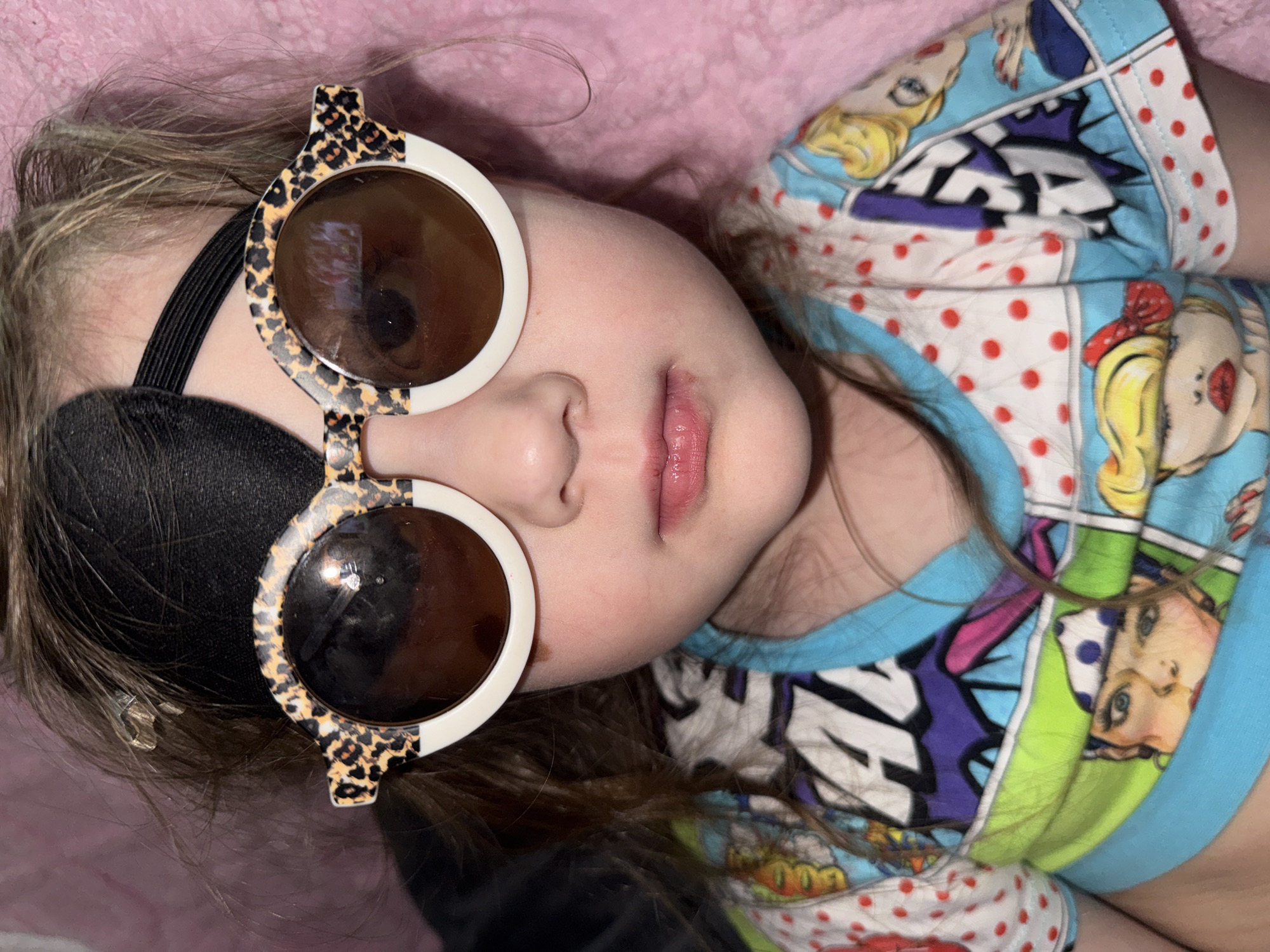 A frontal, waist-up portrait captures a young child located in Кипчак, Russia. The child has light brown, disheveled hair, with a small metallic hair clip visible on the right side of the head. A black eye patch is worn over the left eye, secured by a black elastic strap around the head. Round sunglasses, featuring a white frame with a leopard print pattern on the upper half, cover both eyes; the lenses are dark brown and reflective. The child's nose and mouth are visible below the sunglasses, with lips appearing slightly parted, glossy, and exhibiting minor chapping on the lower lip. The child is dressed in a short-sleeved shirt with a light blue collar and sleeves. The main body of the shirt displays multiple cartoon-style female characters in a pop art aesthetic, interspersed with white areas containing red polka dots. The immediate background on the right consists of a textured, soft pink material, while the background on the left is dark and indistinct. The child is looking directly forward.
