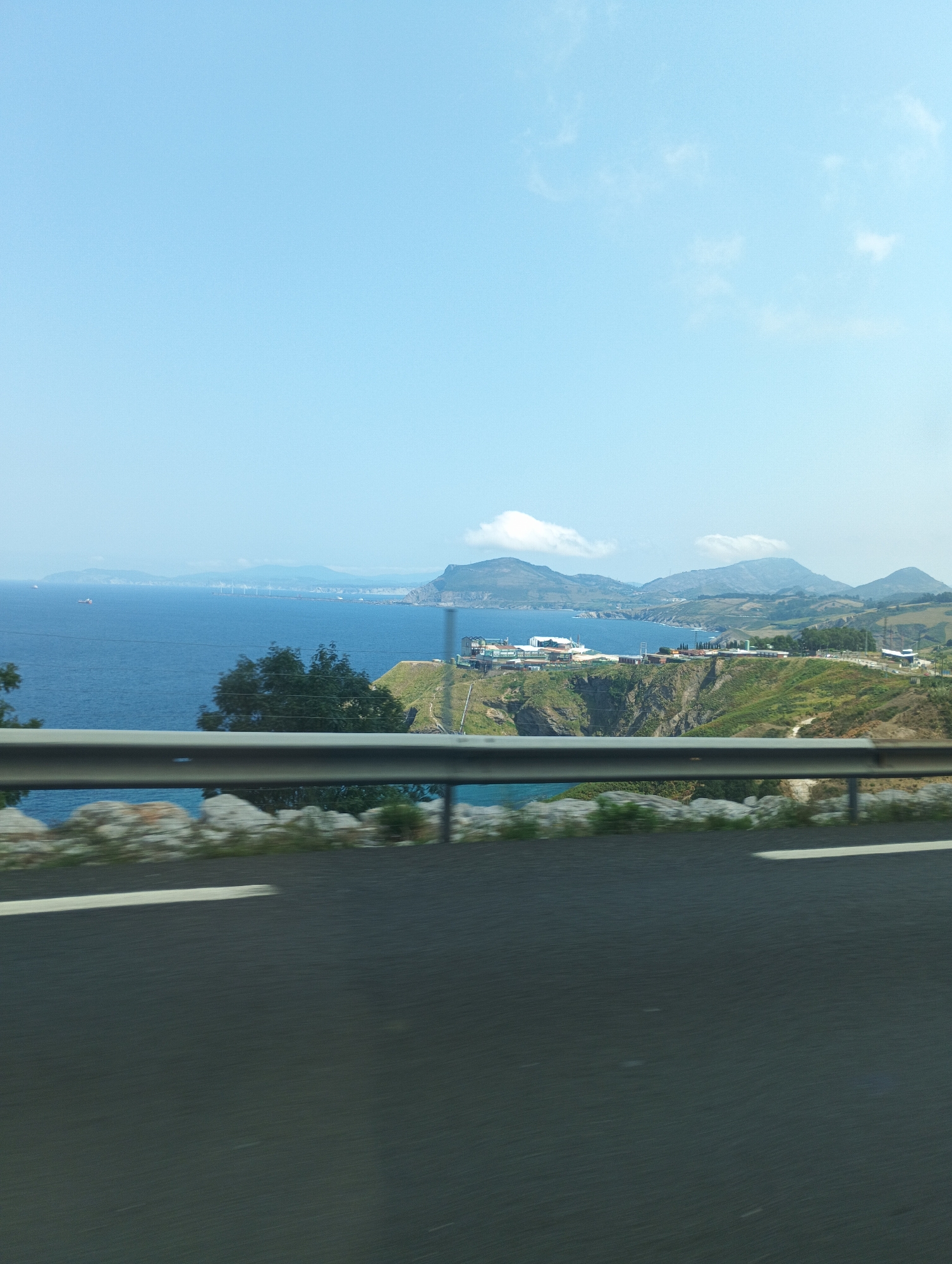 The image shows a coastal landscape viewed from a moving vehicle, likely on a highway. In the foreground, a section of dark asphalt road with a white lane marking is visible, blurred due to motion. A metal guardrail separates the road from a rocky embankment with green vegetation.
Beyond the embankment, a vast expanse of blue ocean stretches to the horizon. The water appears calm. In the middle ground, a rugged coastline with steep cliffs is prominent. Buildings, possibly industrial or residential, are clustered on the top of these cliffs, with some infrastructure like pipelines visible. Green hills and mountains rise behind the coastline, fading into the distance under a clear blue sky with a few scattered clouds. A few ships and wind turbines can be seen on the water and land in the background, suggesting a populated and industrialized coastal area. The overall impression is a scenic drive along a coastline on a bright, sunny day.