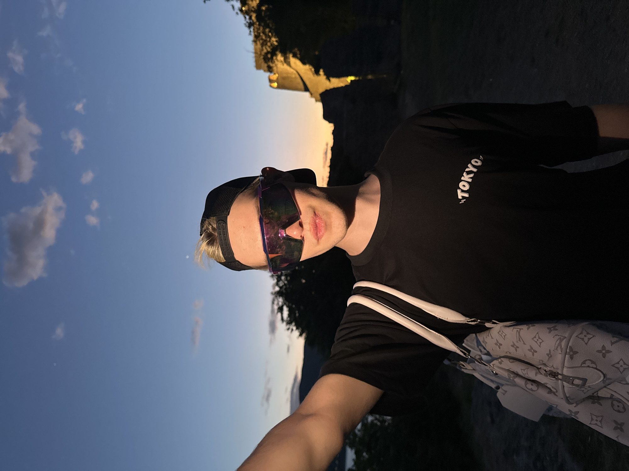 A young person wearing a black shirt, a black baseball cap, and reflective sunglasses is standing outside in Herceg Novi, Montenegro. The shirt has the word "TOKYO" written on it. A white Louis Vuitton duffel bag is visible hanging from their shoulder. The background shows a twilight sky and some buildings on a hill.