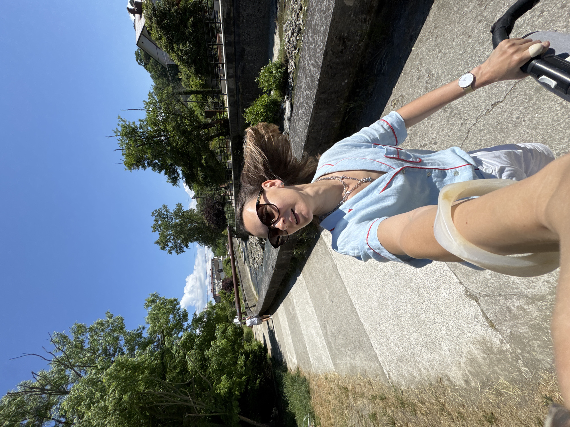 A woman is captured in a first-person perspective selfie, holding a stroller while walking on a paved path alongside a dried-up riverbed. She is wearing a light blue pajama-style top with red trim and white shorts. Large, dark sunglasses are perched on her nose, and her hair is being blown by the wind. A silver necklace with a pendant is visible around her neck. The path is bordered by dry grass on one side and a concrete embankment with rocks and sparse vegetation on the other. In the background, trees and buildings are visible, suggesting an urban or semi-urban setting. The sky is clear and blue, indicating a sunny day. The scene is likely located in Sochi, Russia, given the mention of the city in the prompt.