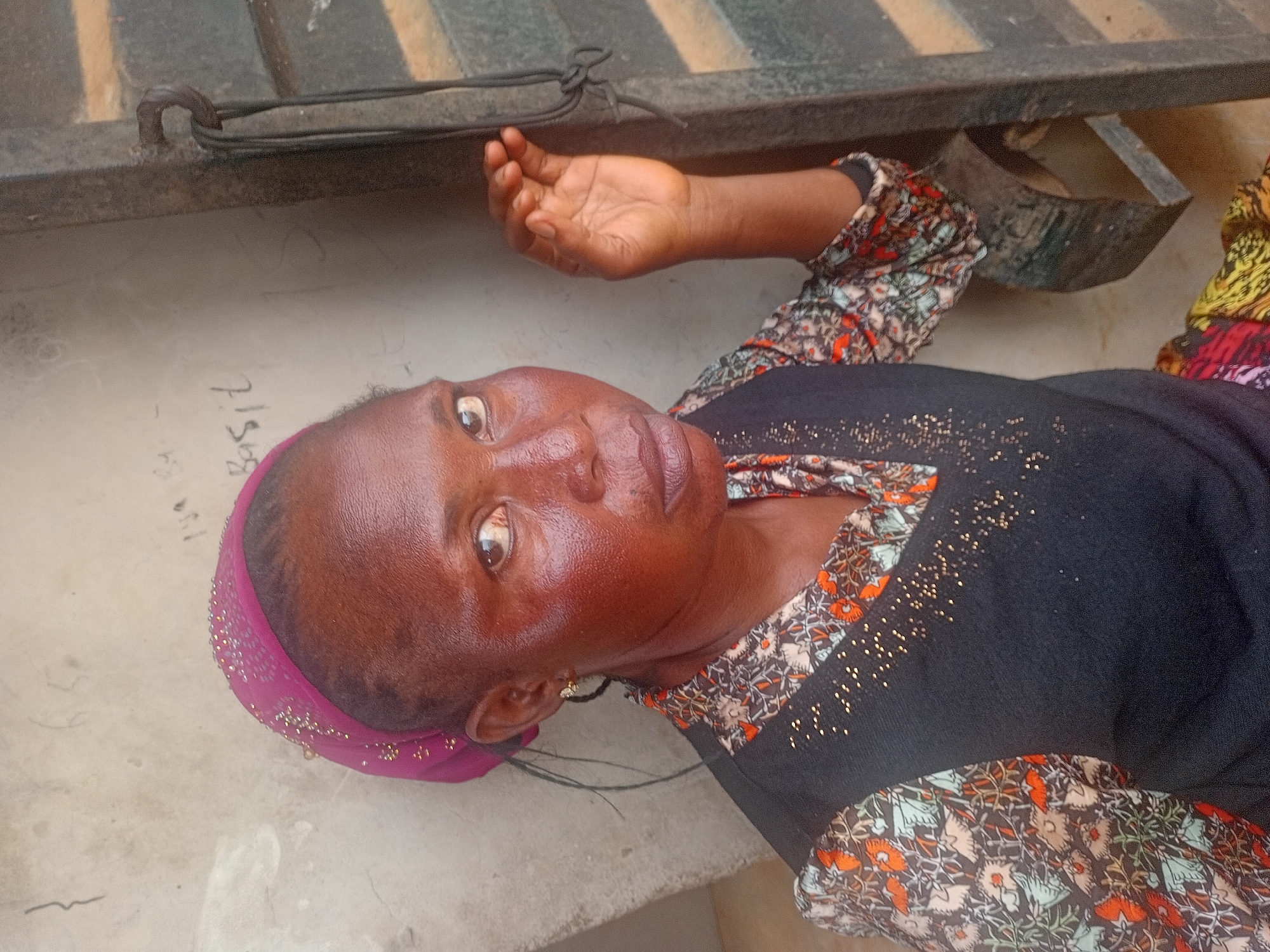 A woman lies on her back on a gray surface beneath a dark metal structure. She is wearing a dark sweater with floral print sleeves and a pink head covering. Her right arm is outstretched. The location is likely Ikorodu, Nigeria, but there's no evidence of this. The word "Basil" is written on the surface to her right.