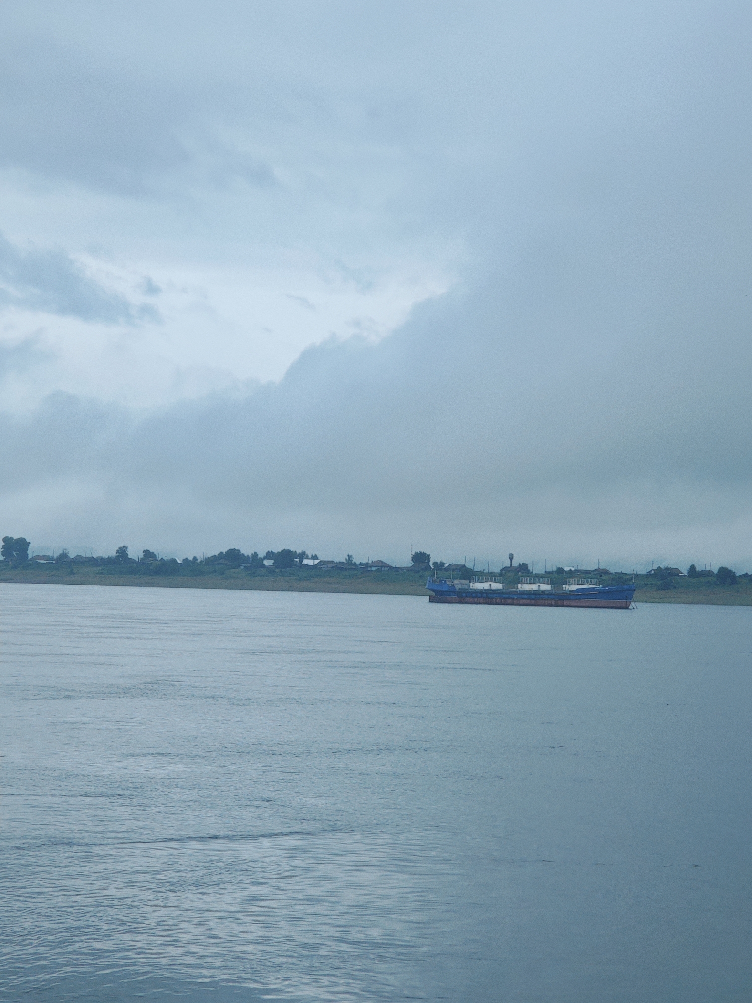 A long, blue and red barge is moored on a wide, calm river. The river takes up the lower half of the image. On the far bank, a line of trees and buildings suggests a settlement. Above this, a thick, grey overcast sky dominates the upper half of the frame. The scene appears to be set in the city of Lesosibirsk, Russia.