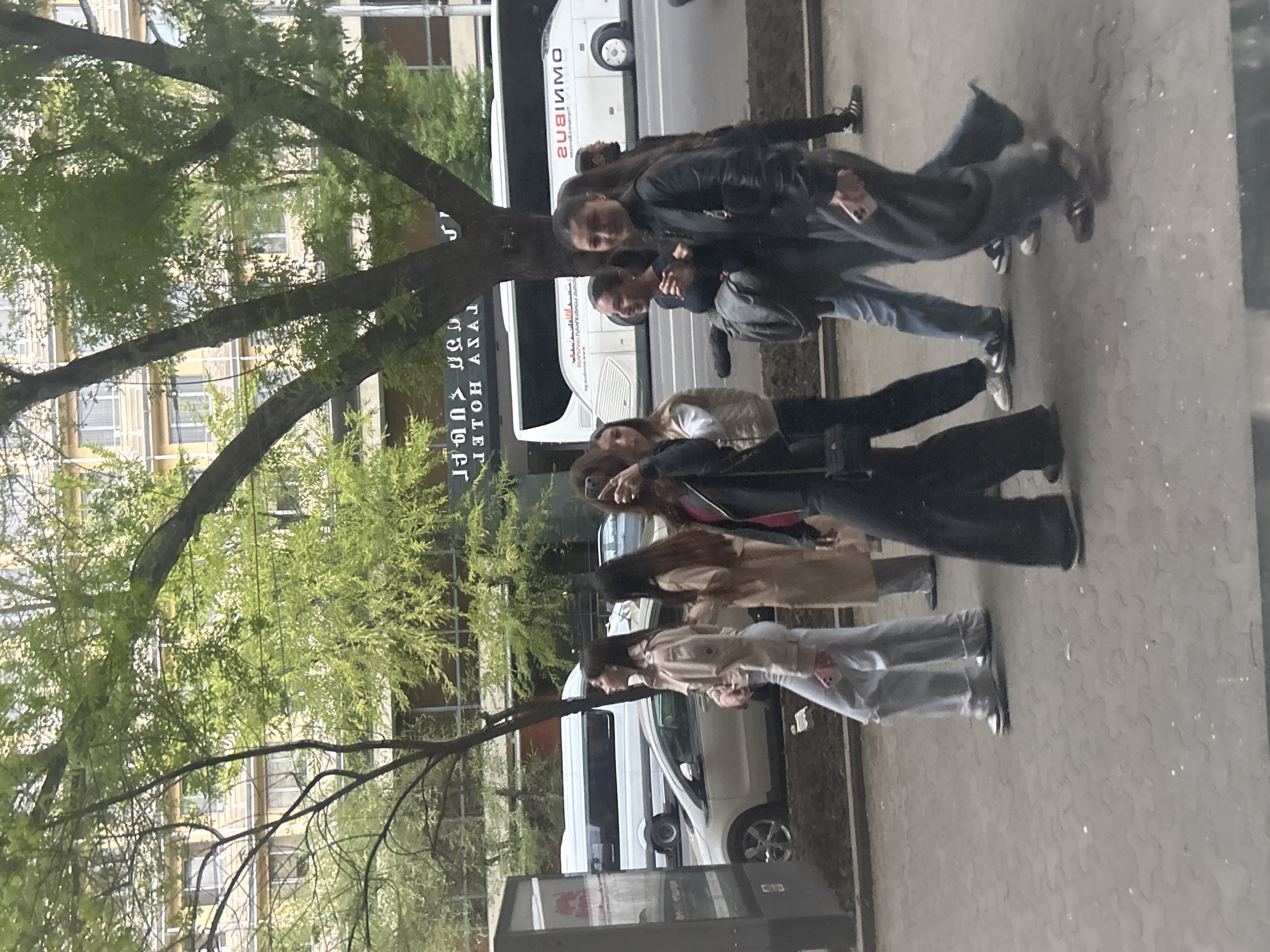 The image depicts an urban street scene in Yerevan, Armenia, under what appears to be an overcast sky during daytime. A group of approximately six young individuals, likely teenagers or young adults, are seen walking along a paved sidewalk. They are dressed in contemporary casual clothing, including jackets, jeans, and wide-leg trousers, with some holding mobile phones.
A large tree with green foliage occupies the left side of the frame, its branches extending across the view and partially obscuring the background. To the right of the tree and on the street, a large white bus is prominently visible. The bus features mirrored English text "OMNIMMO" and "TOUR AND TRANSFER," alongside its Armenian equivalent "ՕՄՆԻՄՈՒ." Further down the street, another white car and a dark vehicle can be seen.
Behind the vehicles and through the foliage, multi-story buildings with numerous windows are visible, suggesting a commercial or residential urban area. A dark rectangular sign, also with mirrored text, reads "OMNIMOU" and "TOUR HOTEL," accompanied by Armenian script "ՕՄՆԻՄՈՒ ՀՈՒՌՀԵՌ." The sidewalk is paved with light grey blocks, indicating a well-maintained public space. The overall impression is a bustling street environment captured during a typical day.