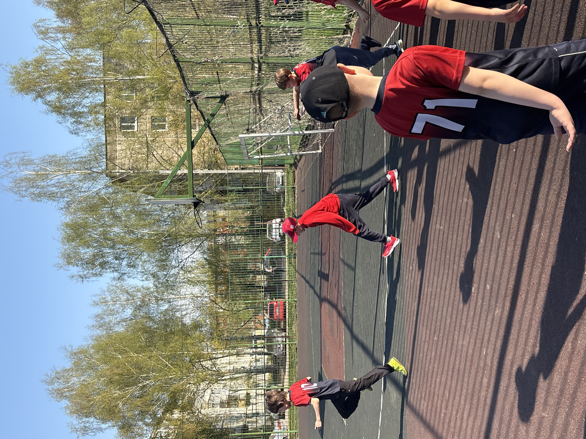 The image shows a group of children outdoors, likely on a sports field or playground in Izhevsk, Russia. They appear to be engaged in some kind of physical activity or exercise routine. The children are wearing athletic clothing. The background includes trees, a fence, and a building that is likely a residential building. The sky is blue and the image appears to have been taken on a sunny day.