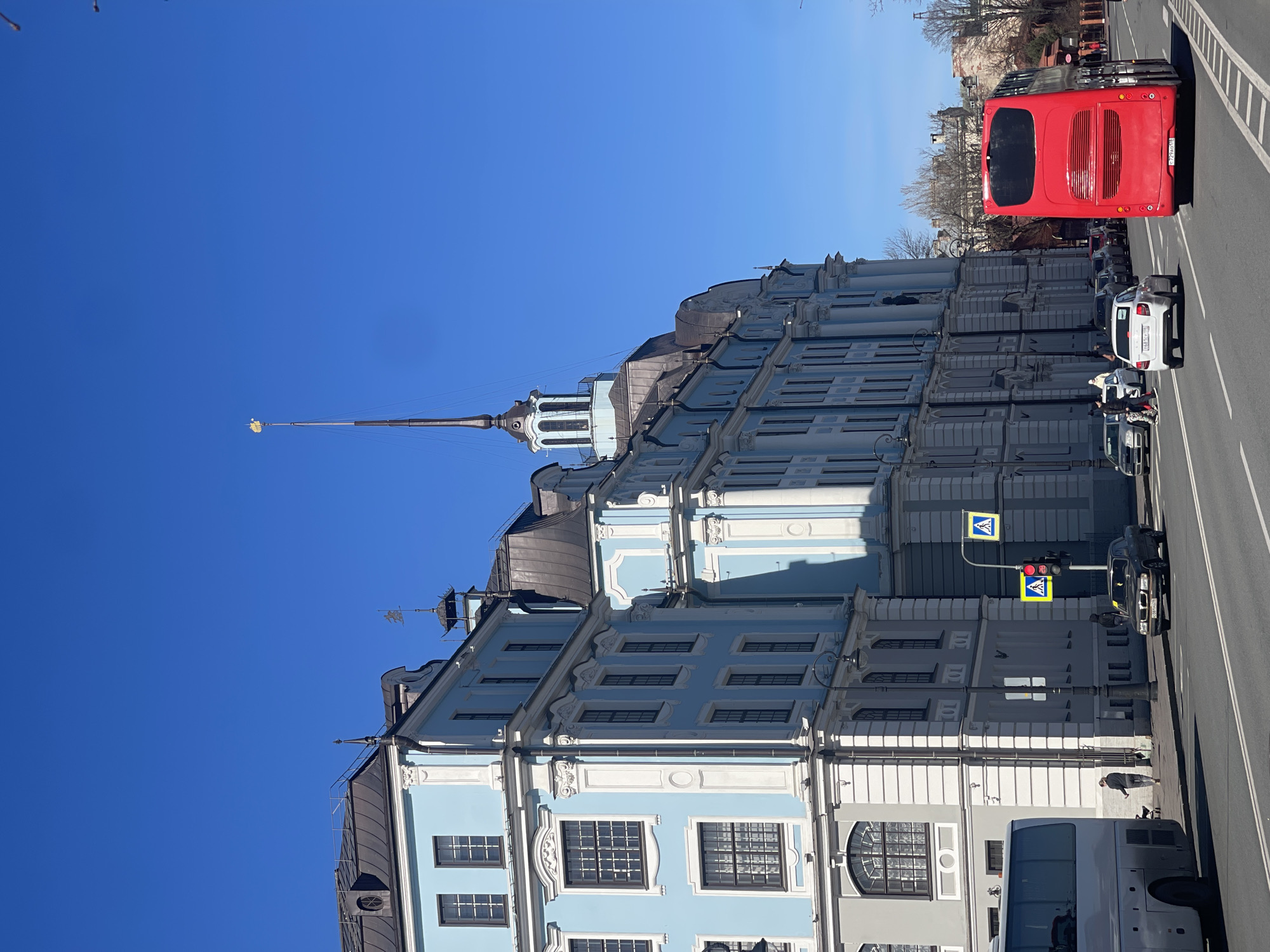 The image shows a light blue and white multi-story building in Saint Petersburg, Russia, with a tall spire topped with a golden weather vane. The building stands beside a road with traffic, including a red double-decker bus and several cars. A pedestrian is visible on the sidewalk near a pedestrian crossing sign and a traffic light. The sky is clear and blue.