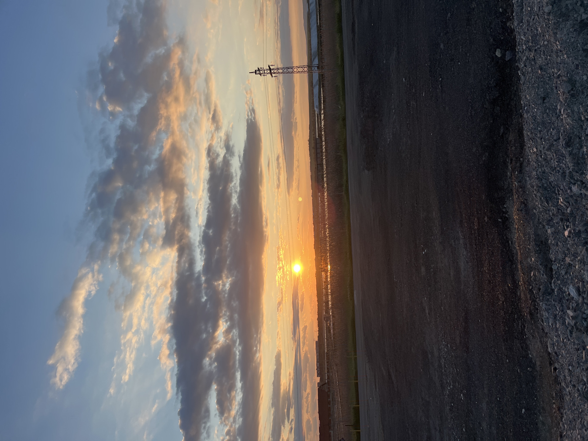The image captures a sunset scene near Burgas, Bulgaria. The sky is filled with streaks of orange and yellow light illuminating clouds in the fading sunlight. A power transmission tower stands out near the horizon. In the foreground, there's a stretch of paved area next to what looks like a gravel or stone shoulder. The overall setting is an industrial or transitional zone on the outskirts of the city.