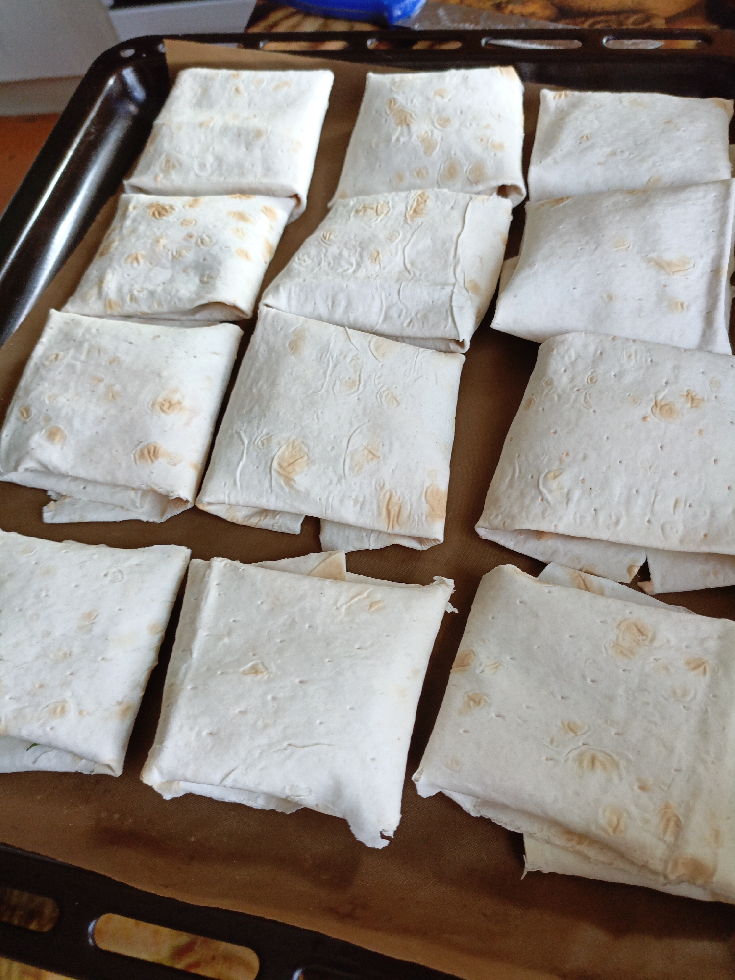 A baking sheet lined with parchment paper contains several square-shaped food items, likely prepared wraps or pastries, arranged in rows. The items are made of thin, light-colored dough, with some pieces showing browning. The baking sheet itself is dark metal with visible handles or edges. The background is blurred but suggests an indoor setting, possibly a kitchen, with some indistinct objects and colors. The image is taken from a top-down perspective. This image is located in Novoznamenskaya 1-Ya, Russia.